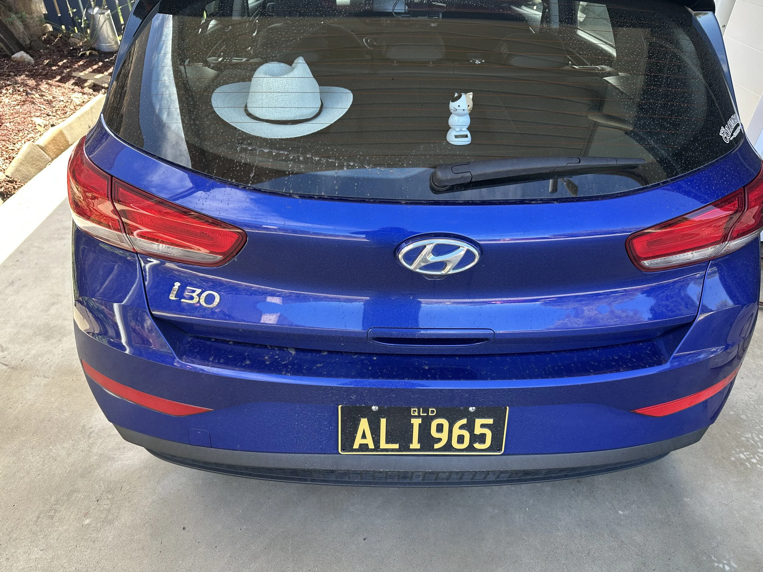 The rear view of a blue Hyundai i30 car with a black and yellow Queensland license plate reading 'AL1965.' On the back window, there is a white cowboy hat and a white figurine of a cat with a black patch on its head.