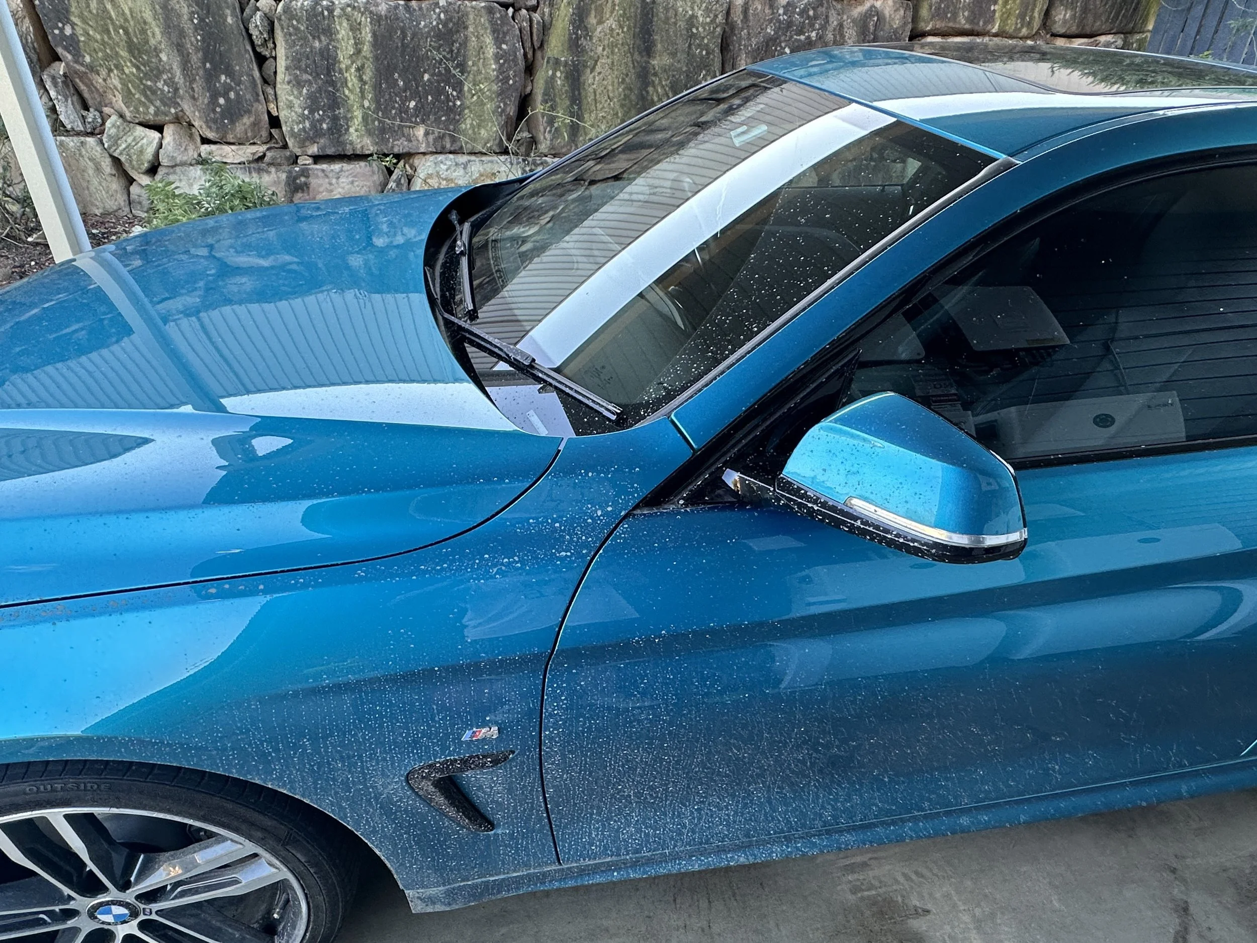 Close-up of front part of a metallic blue BMW car with a section of the front wheel and a stone wall in the background.