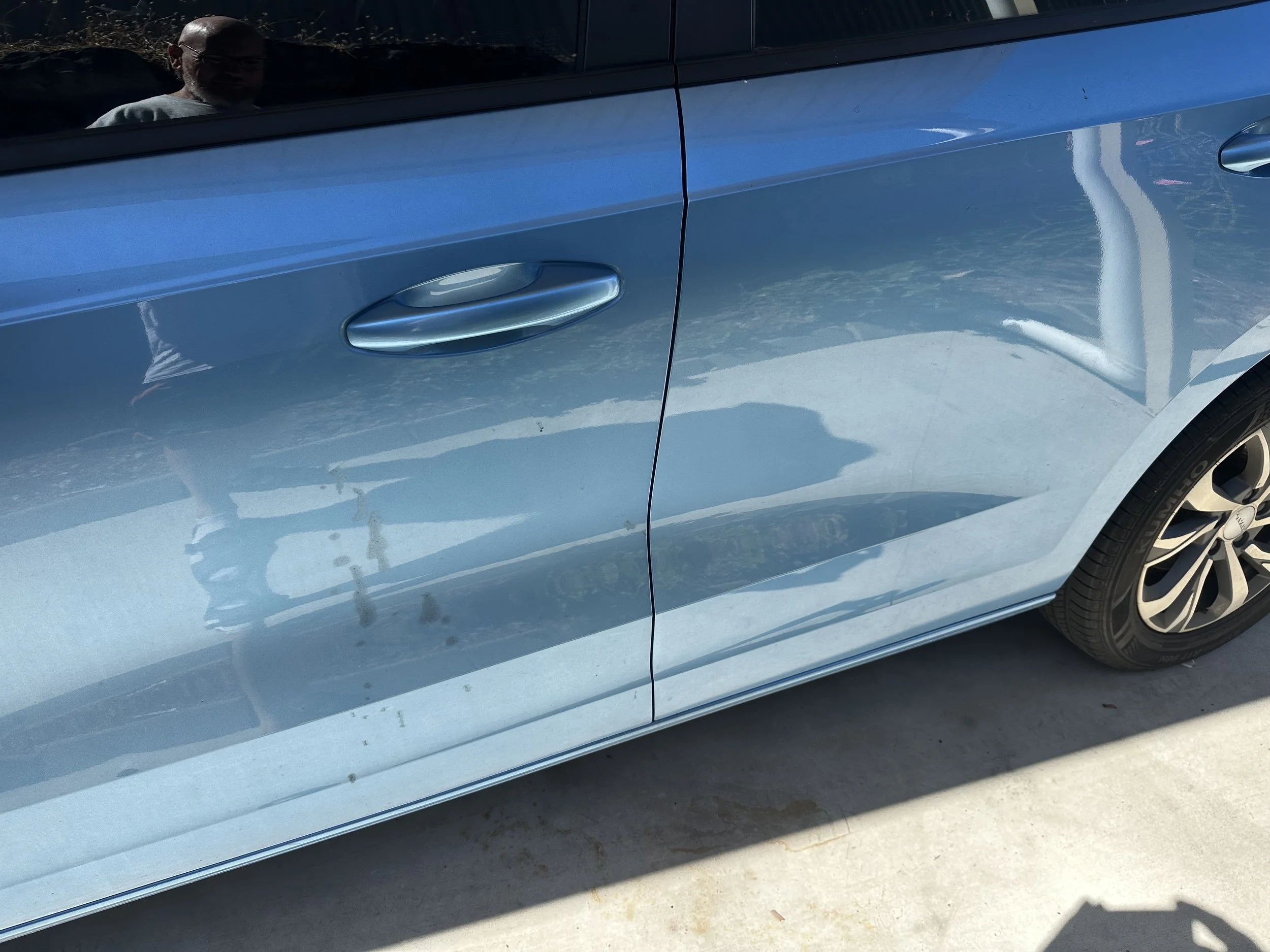 Close-up of a blue car's side, showing the car door handle, a reflection of a person in the window, and part of the wheel.