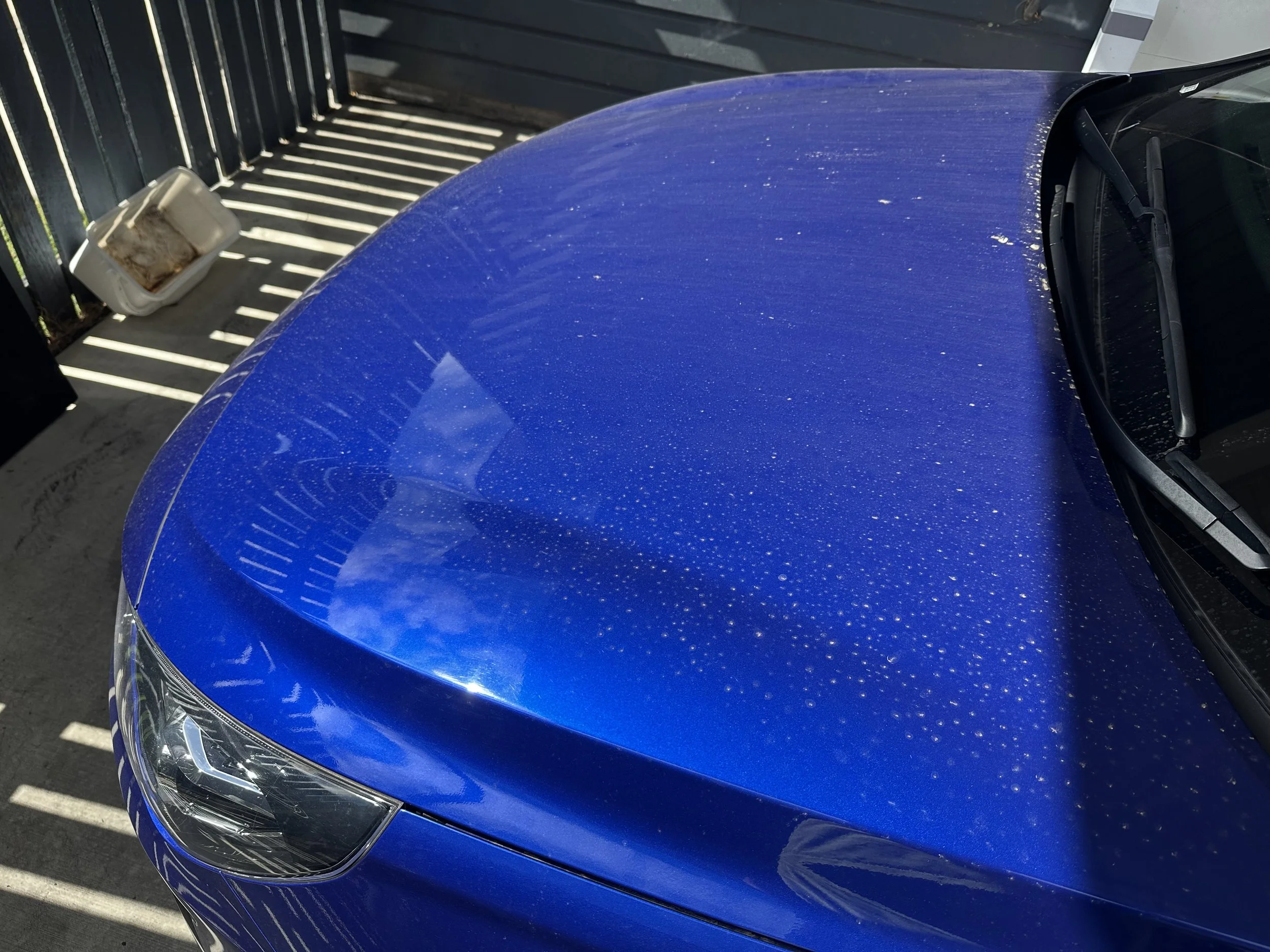 Close-up of a blue car's hood and front windshield, parked in sunlight with shadow patterns from a fence above. A blurred white container with dirt and stains rests on the ground nearby.