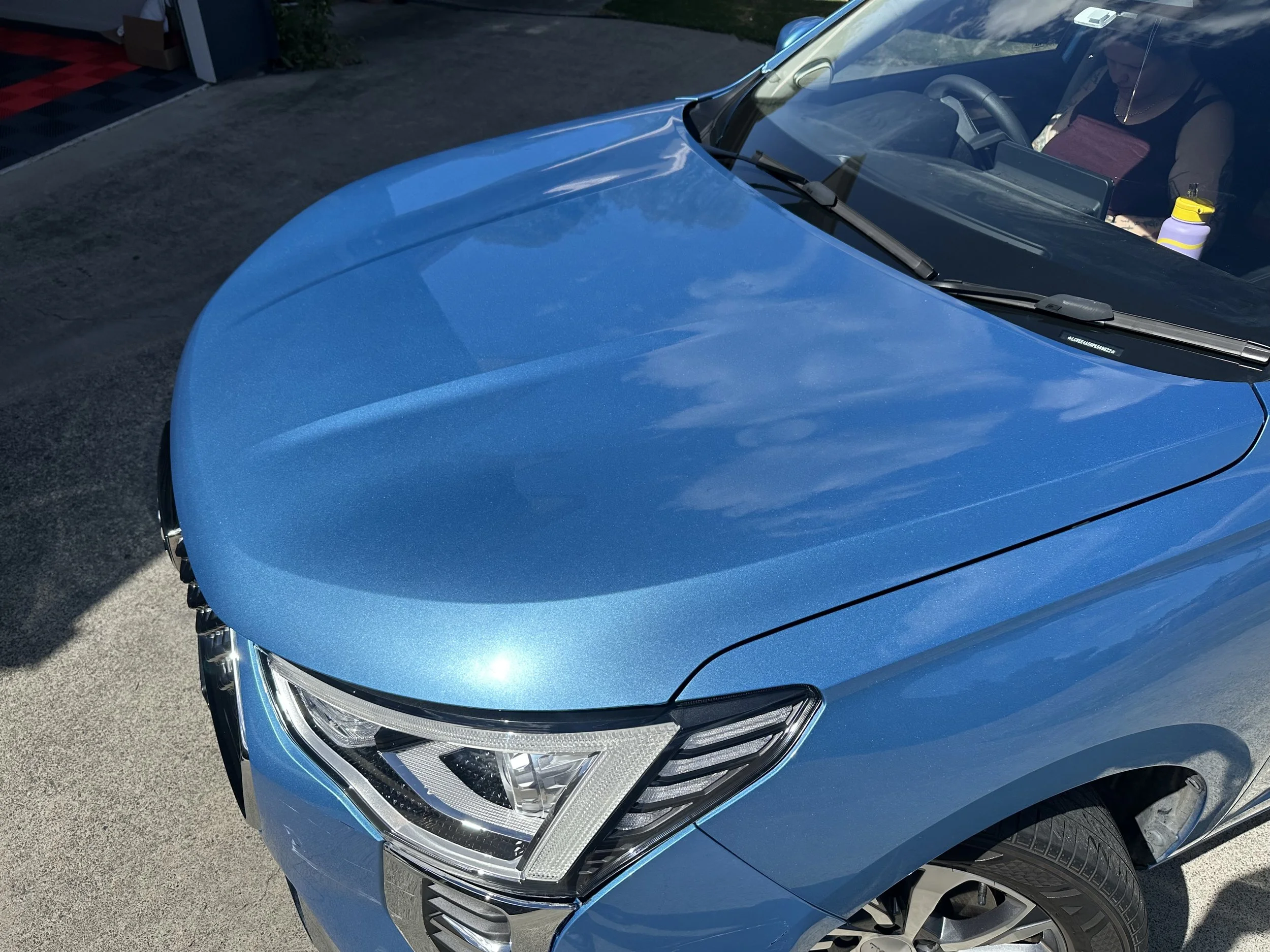 Close-up of the front of a shiny blue car showing the headlight, part of the hood, and windshield, with a person inside and a water bottle on the dashboard.