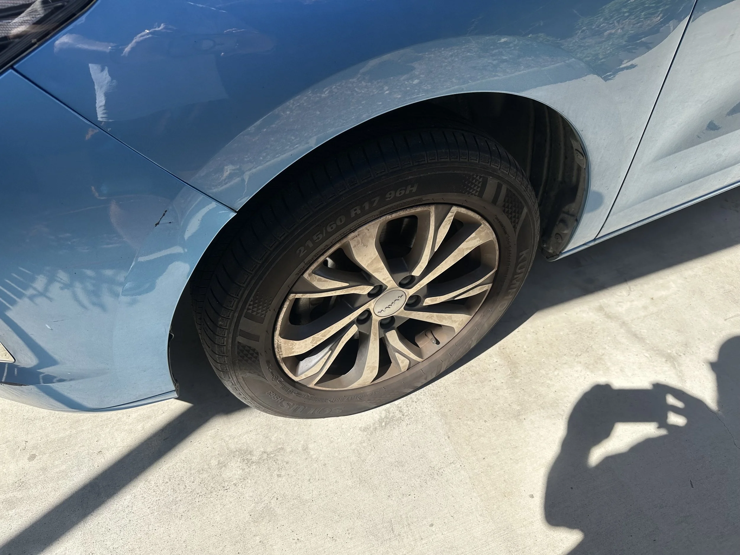 Close-up of a blue car's front tire and wheel on concrete ground with shadows and reflections.