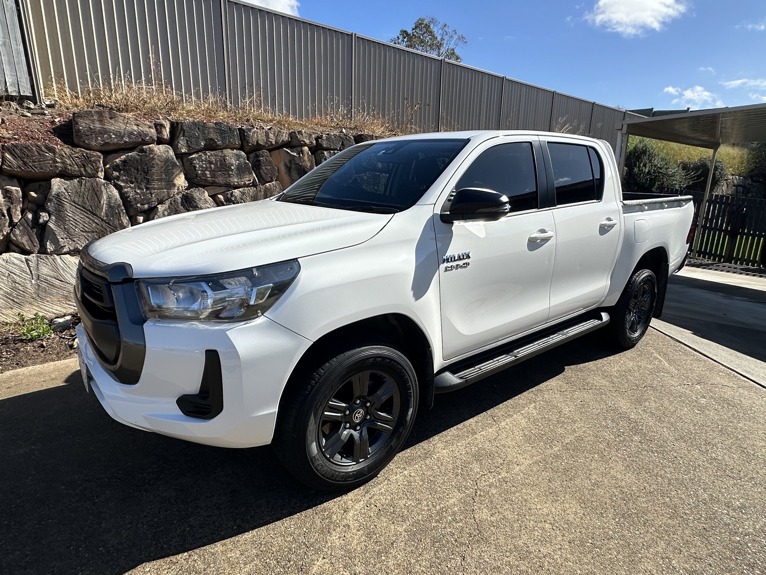 A white pickup truck parked on a concrete surface near a stone retaining wall and metal fence. The truck's windows are tinted and it is under a partly cloudy blue sky.