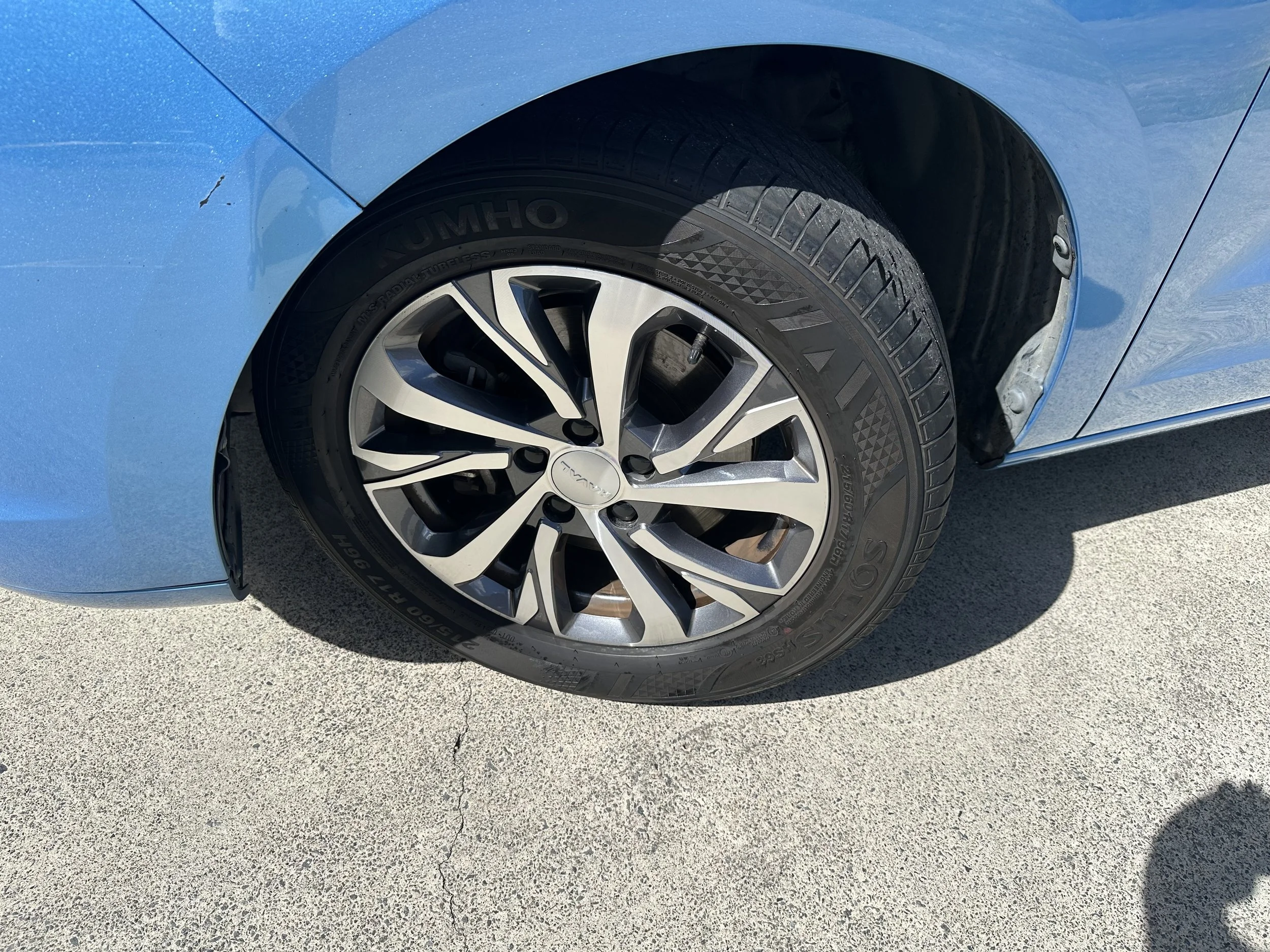 Close-up of a blue car's front wheel and part of the fender, with a black tire and silver rim, parked on a concrete surface.