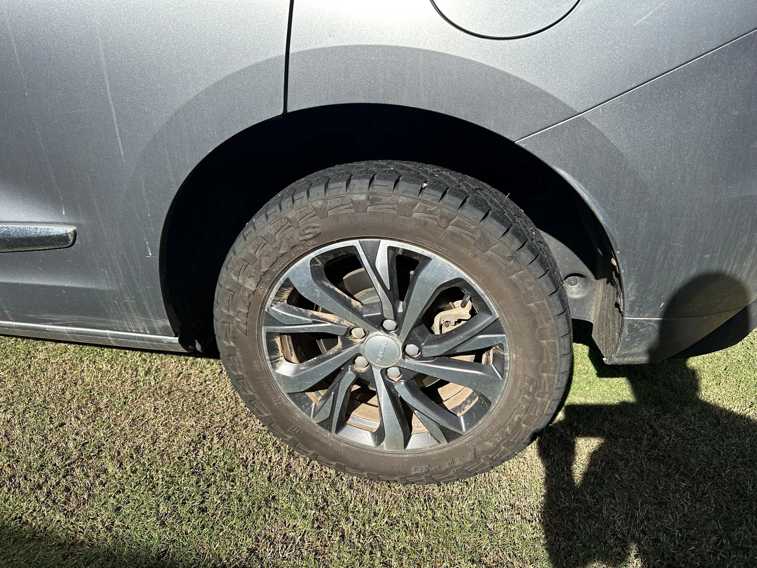 A close-up of a gray car's wheel and tire, with the car parked on grass. The wheel features a multi-spoke design, and the tire has visible tread patterns.