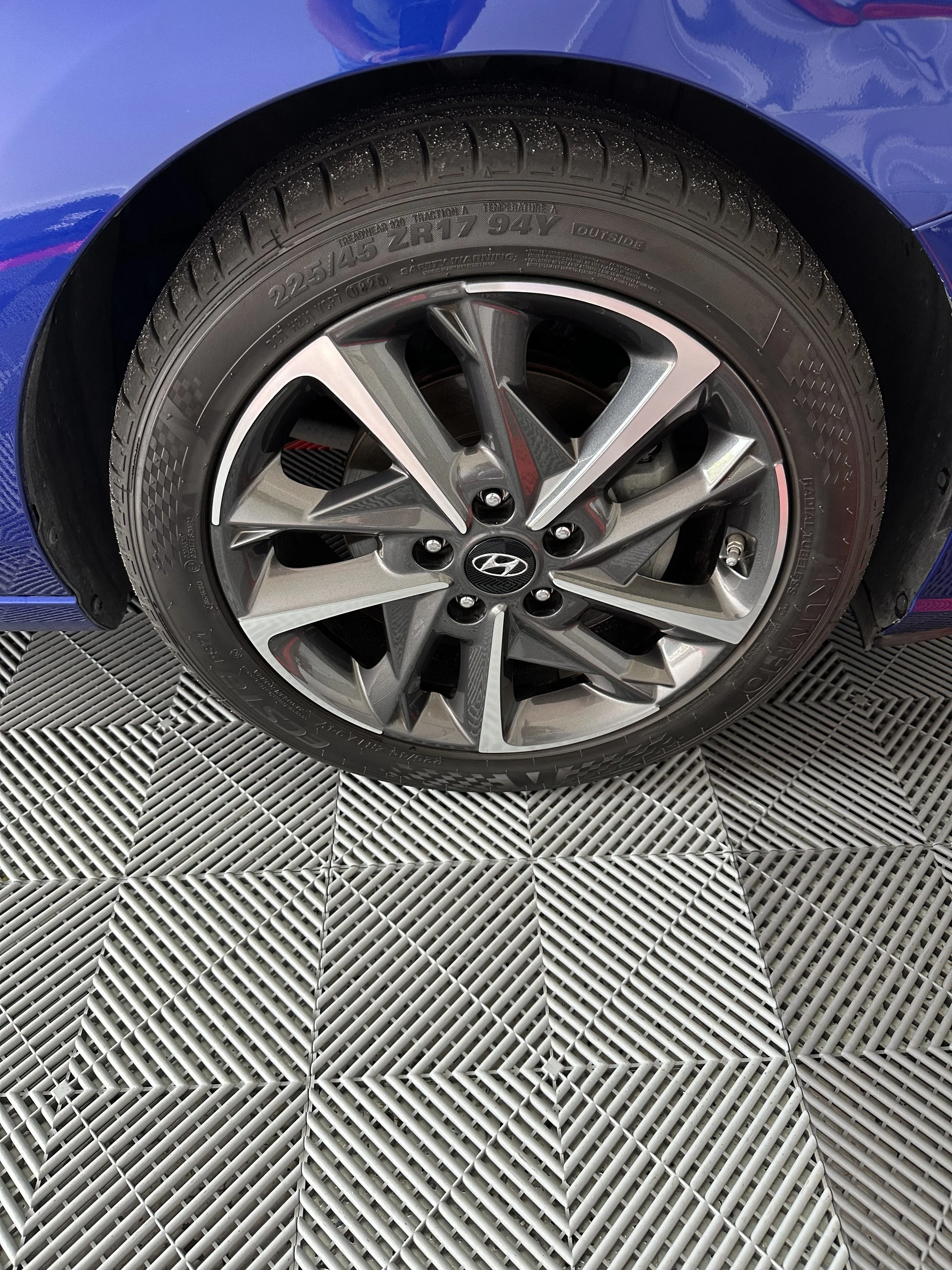 Close-up of a blue car's tire and wheel, with the tire displaying size and specifications, on a textured metal surface.