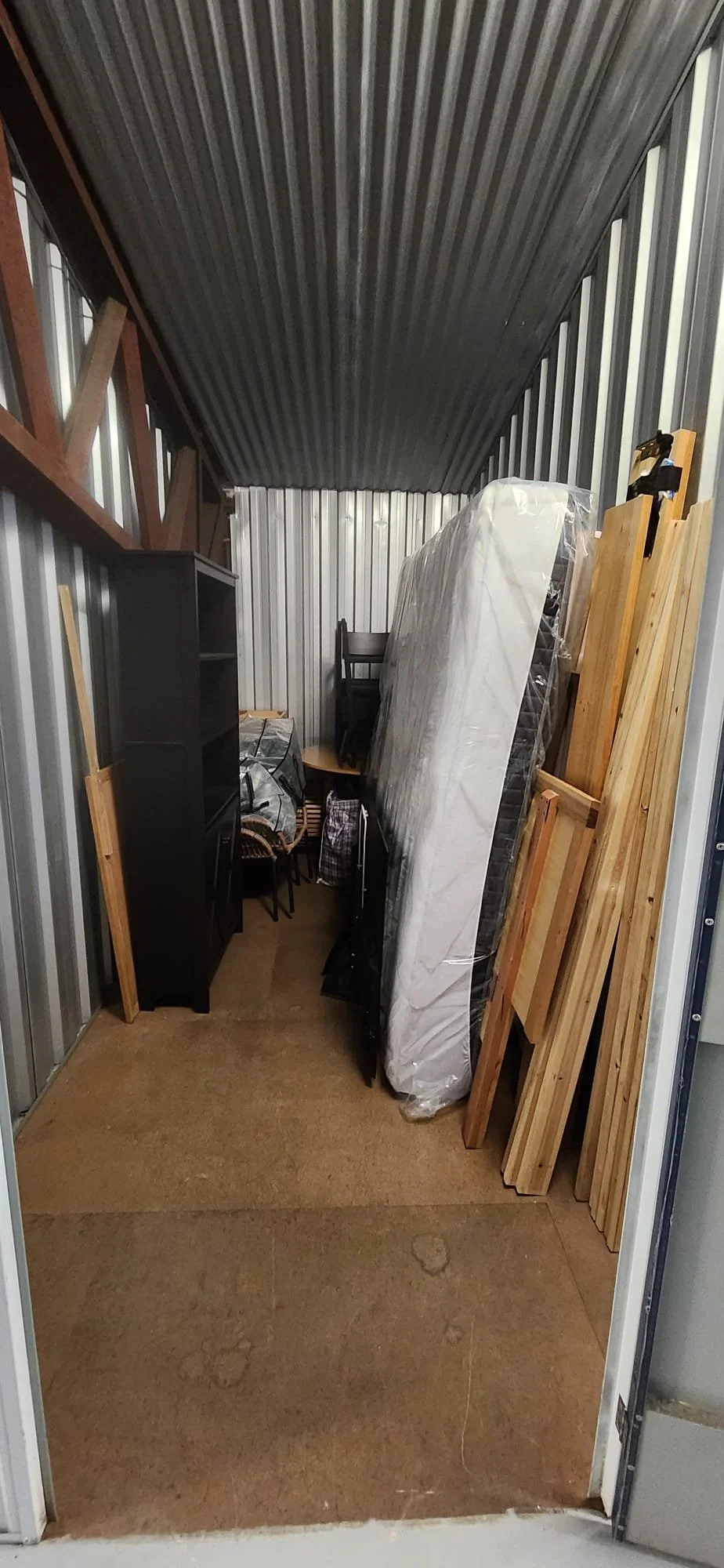 Storage unit filled with furniture and household items, including a mattress in plastic, wooden planks, black bookshelf, chairs, and a small cabinet.