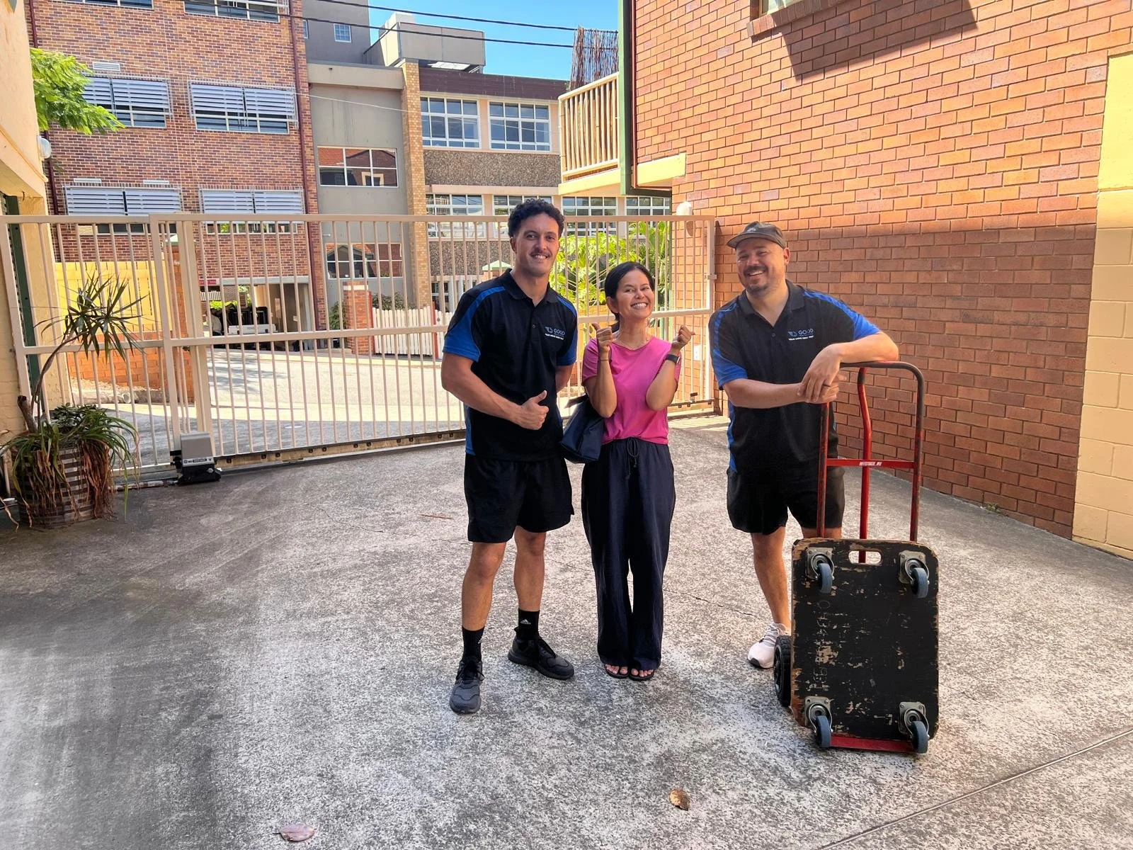 Three people standing outside in front of a gated area along a city street. Two men are wearing black and blue uniforms, and a woman in the middle is wearing a pink shirt and black pants. They are smiling, and the man on the right is resting his arm on a hand truck with a black box on it. Bright sunlight is casting shadows.