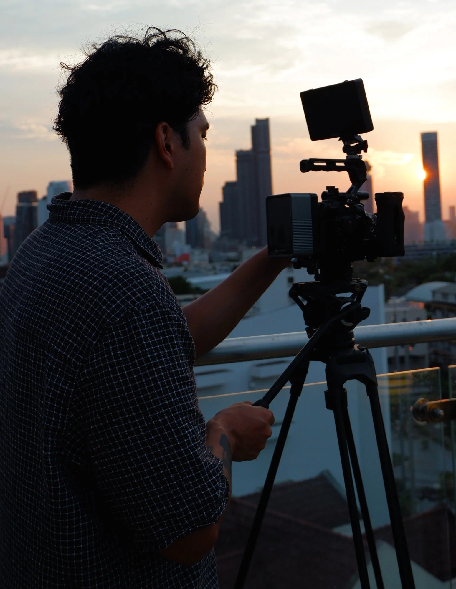 Silhouette of a person operating a camera on a tripod during sunset with a city skyline in the background.
