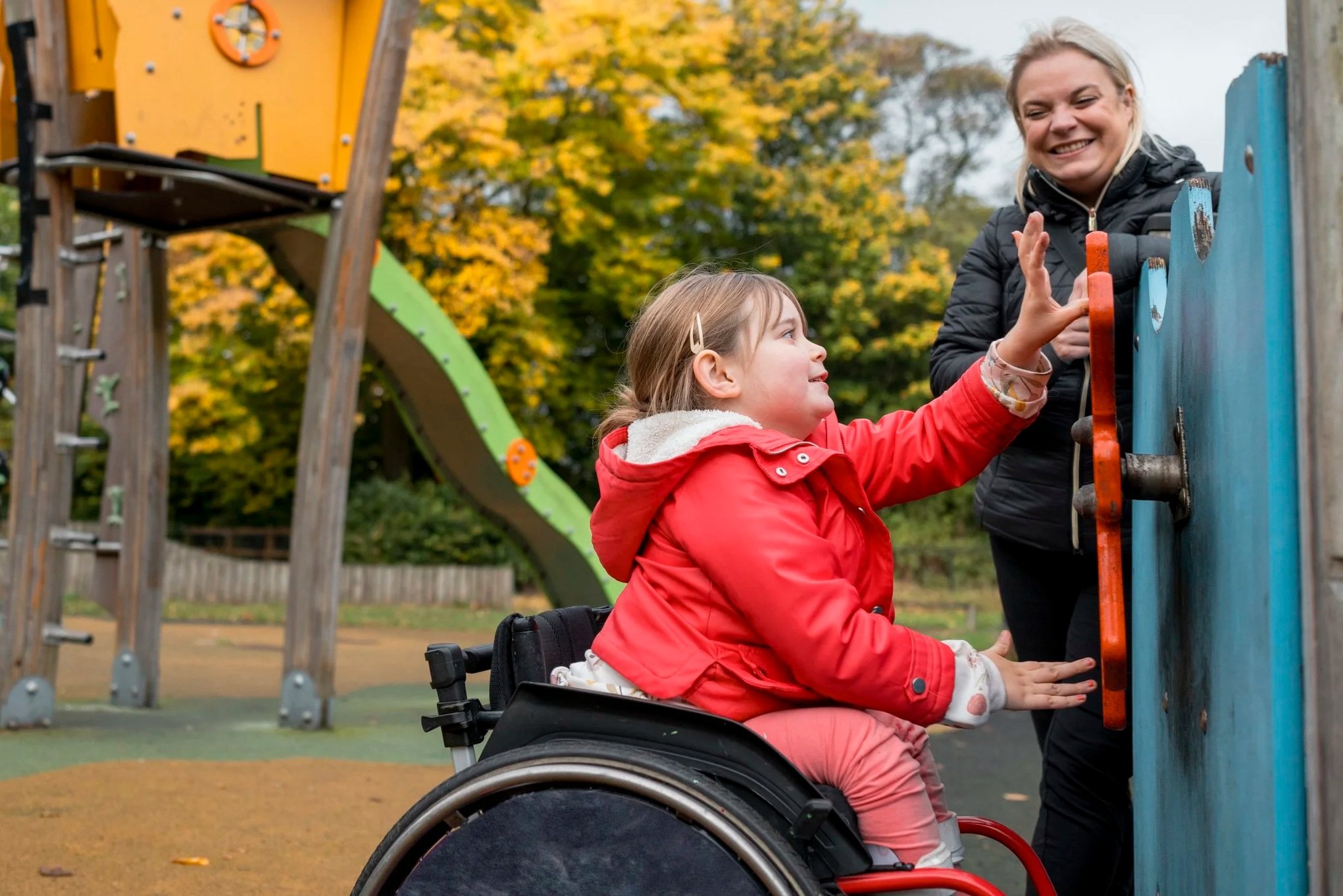 A young girl in a wheelchair reaching up to hold onto a playground equipment while an adult woman assists her. The background shows a playground with a slide and autumn trees.