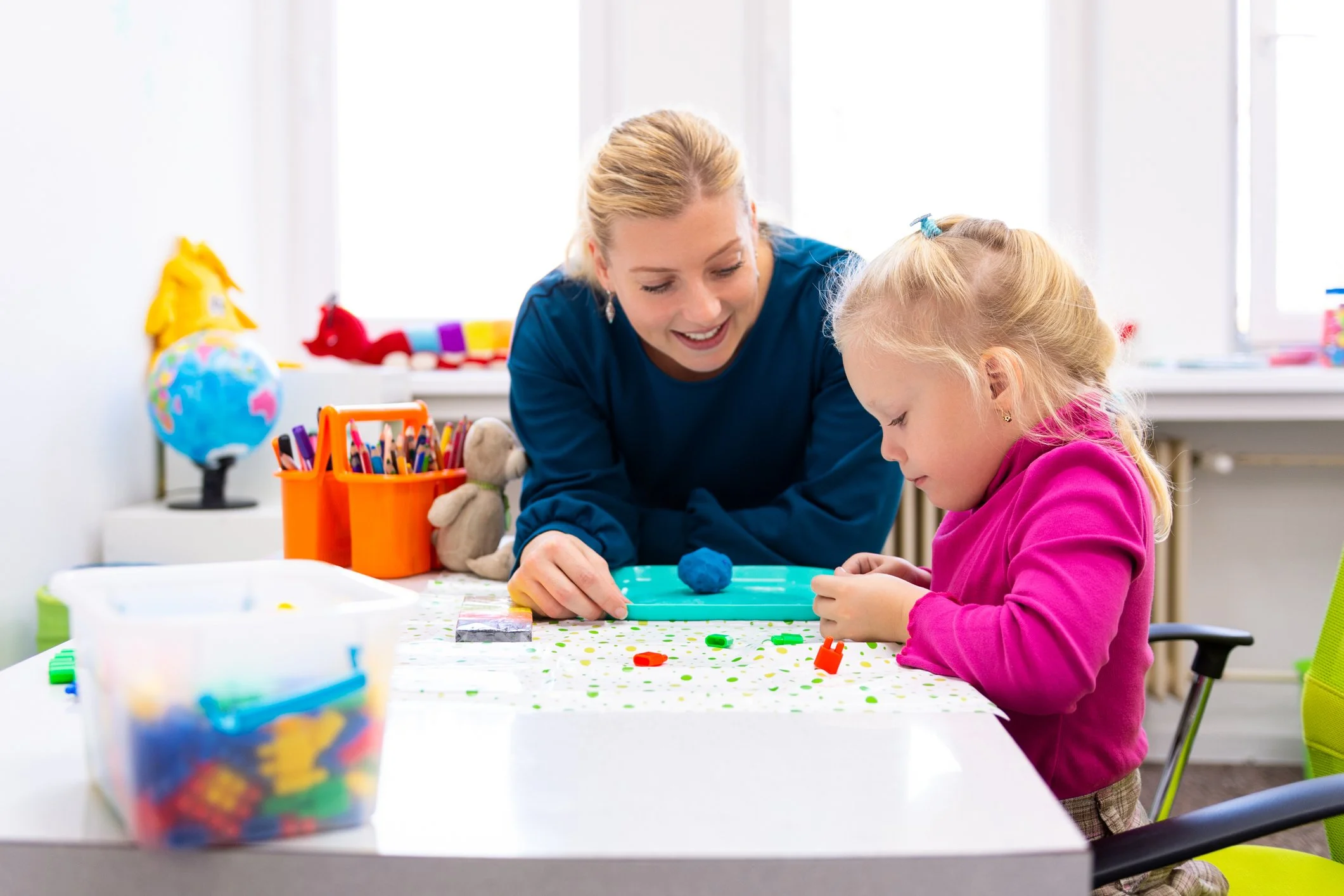 A woman and a young girl sitting at a table, engaging in a craft activity with small colorful objects in a bright room with windows.