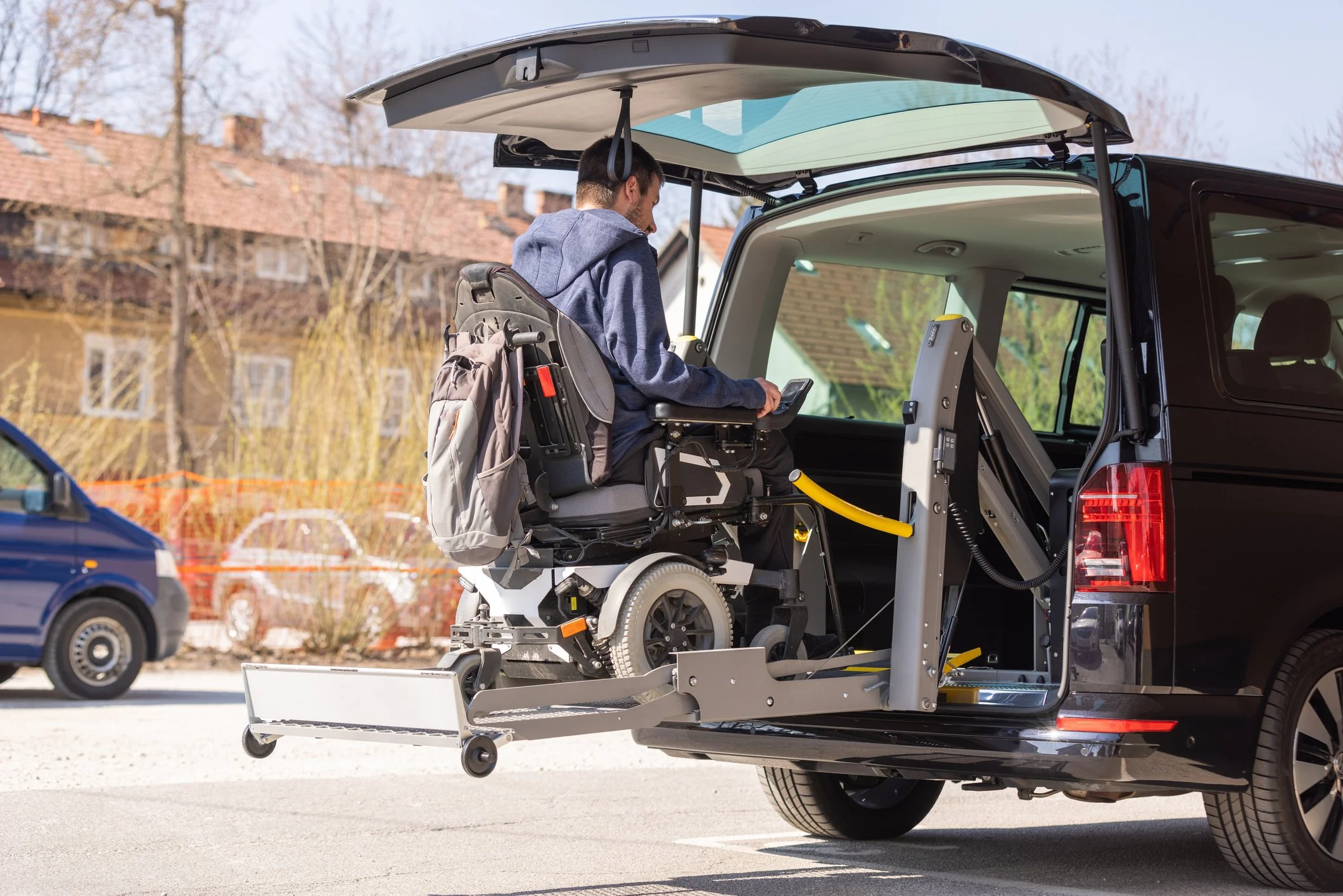 A person in a wheelchair boarding a black van equipped with a lift system for accessibility, parked on a city street with residential buildings and trees in the background.
