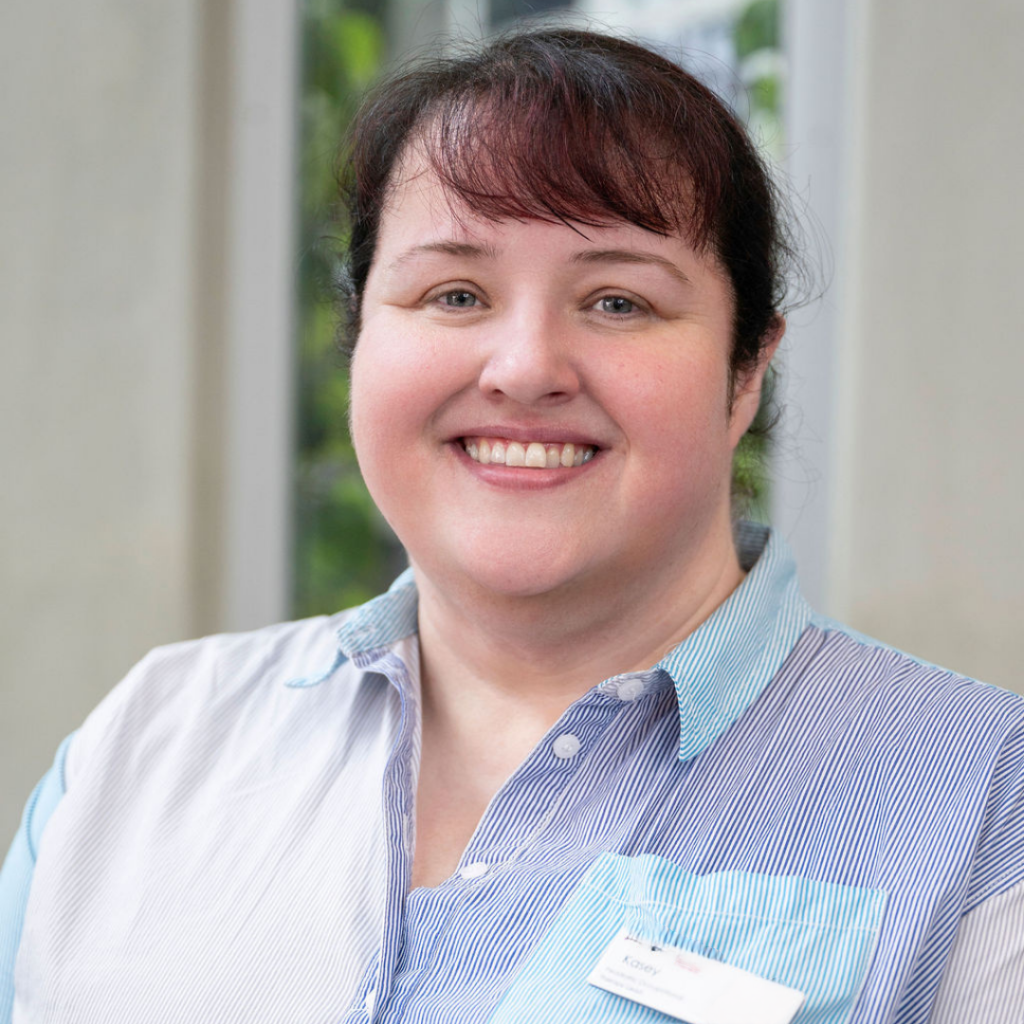 Close-up of a woman with short dark red hair and light skin smiling indoors with a window with greenery in the background.