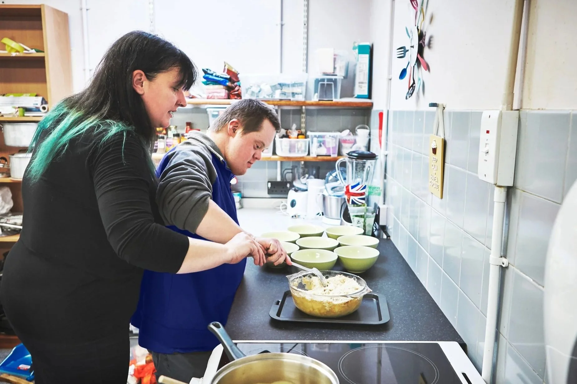 A woman and a man are preparing food together in a kitchen. The woman is stirring a bowl of ingredients while the man looks on, with several empty bowls lined up on the counter.