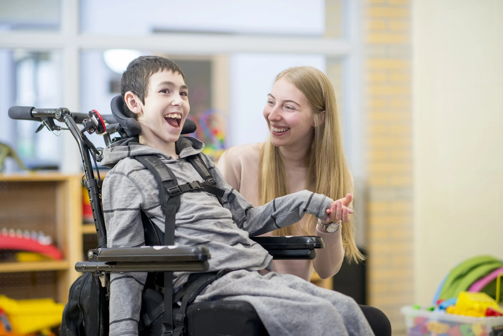 A young boy in a wheelchair smiling and holding hands with a woman who is sitting beside him, in a brightly lit indoor setting.