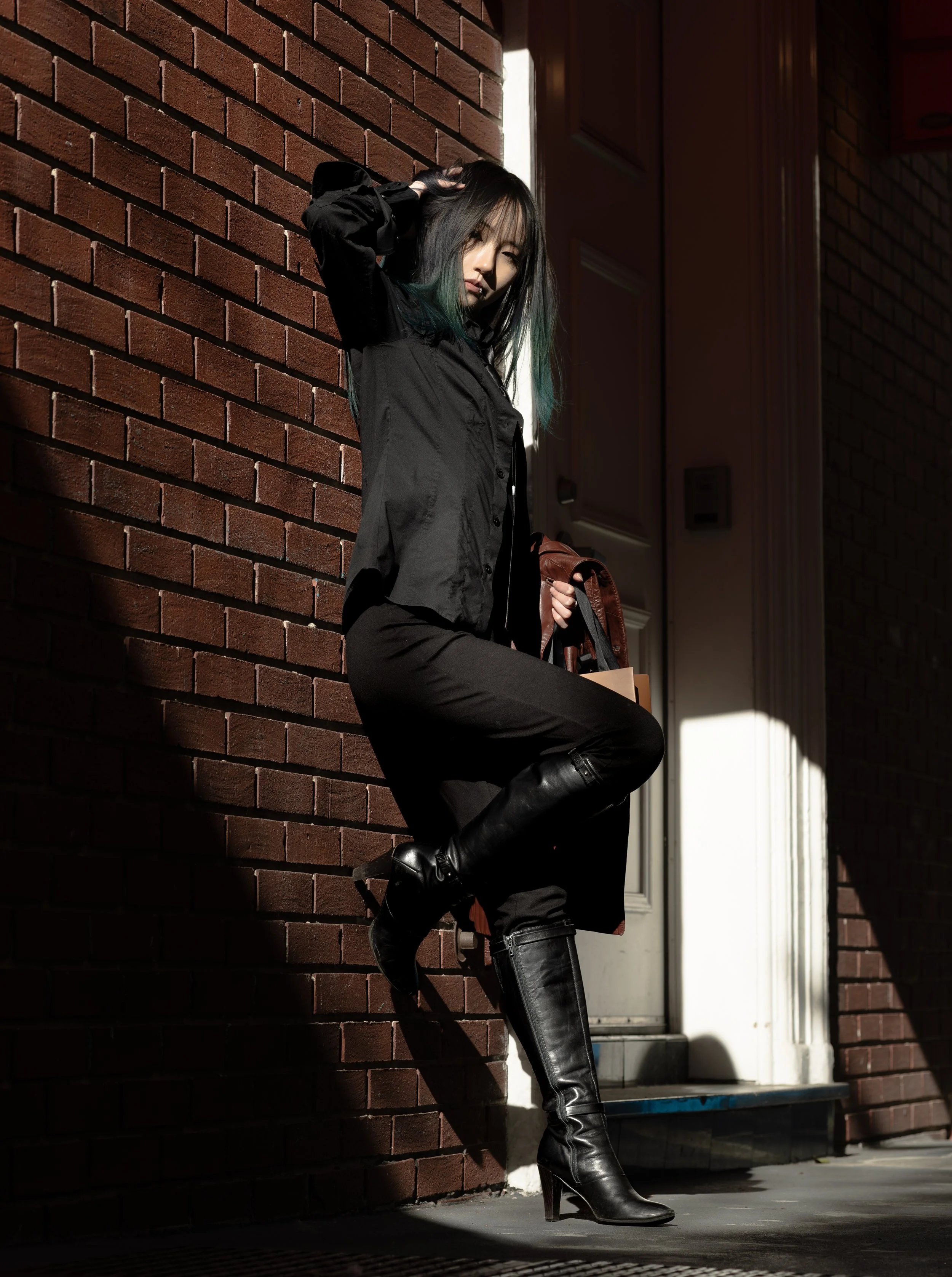 A young woman with dark hair and green tips, wearing a black jacket, black pants, and tall black leather boots, leaning against a brick wall outdoors in sunlight, holding a brown handbag.