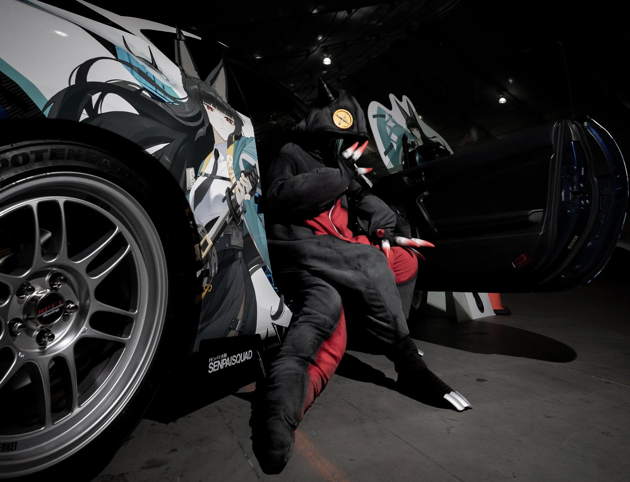 A person dressed in a black costume with a cat-like mask and claws, sitting in a luxury car, at an indoor exhibition or event, with anime-style posters on display behind them.