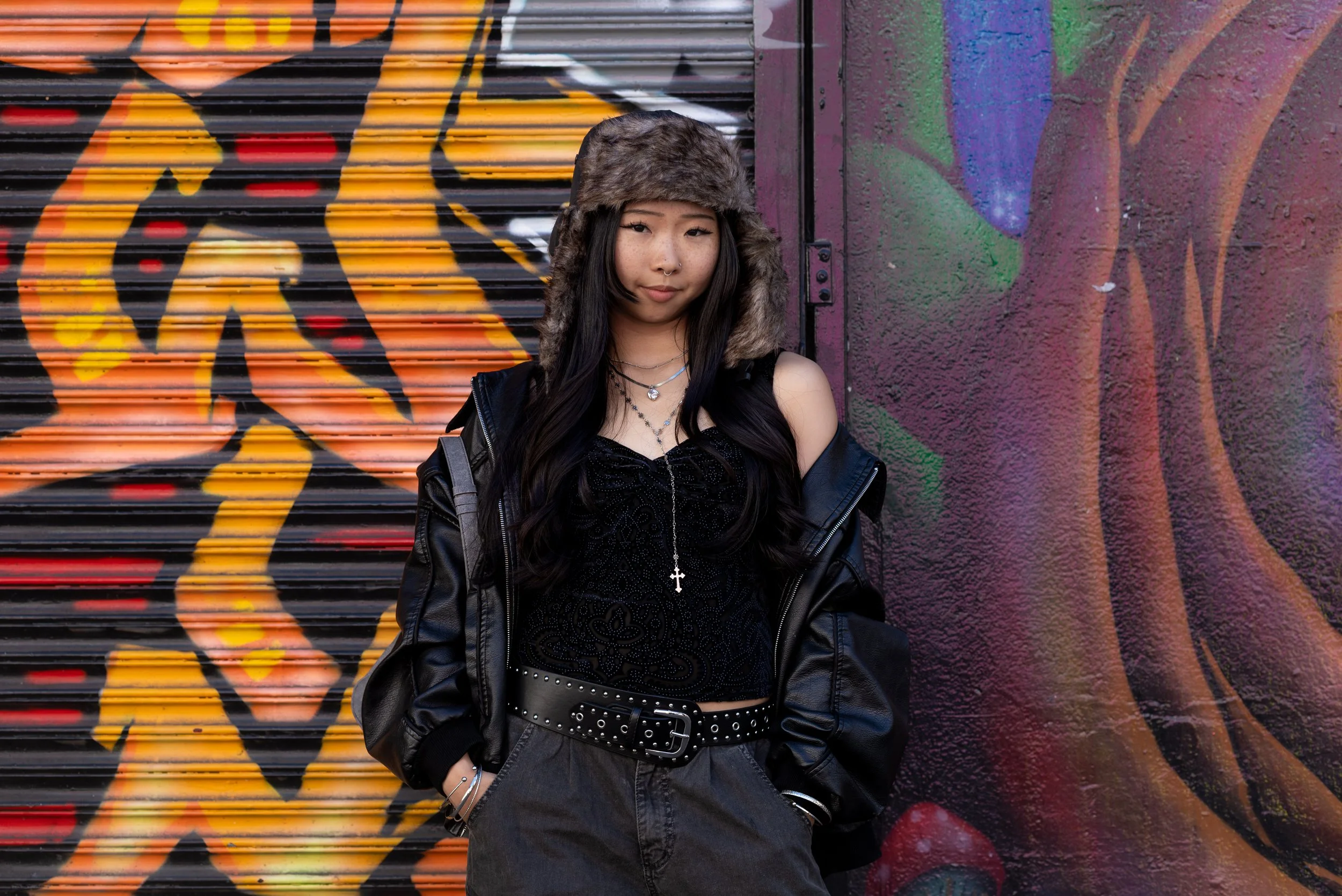 A young woman with long black hair, wearing a fur hat, black lace top, leather jacket, and layered necklaces, standing in front of a colorful graffiti wall.