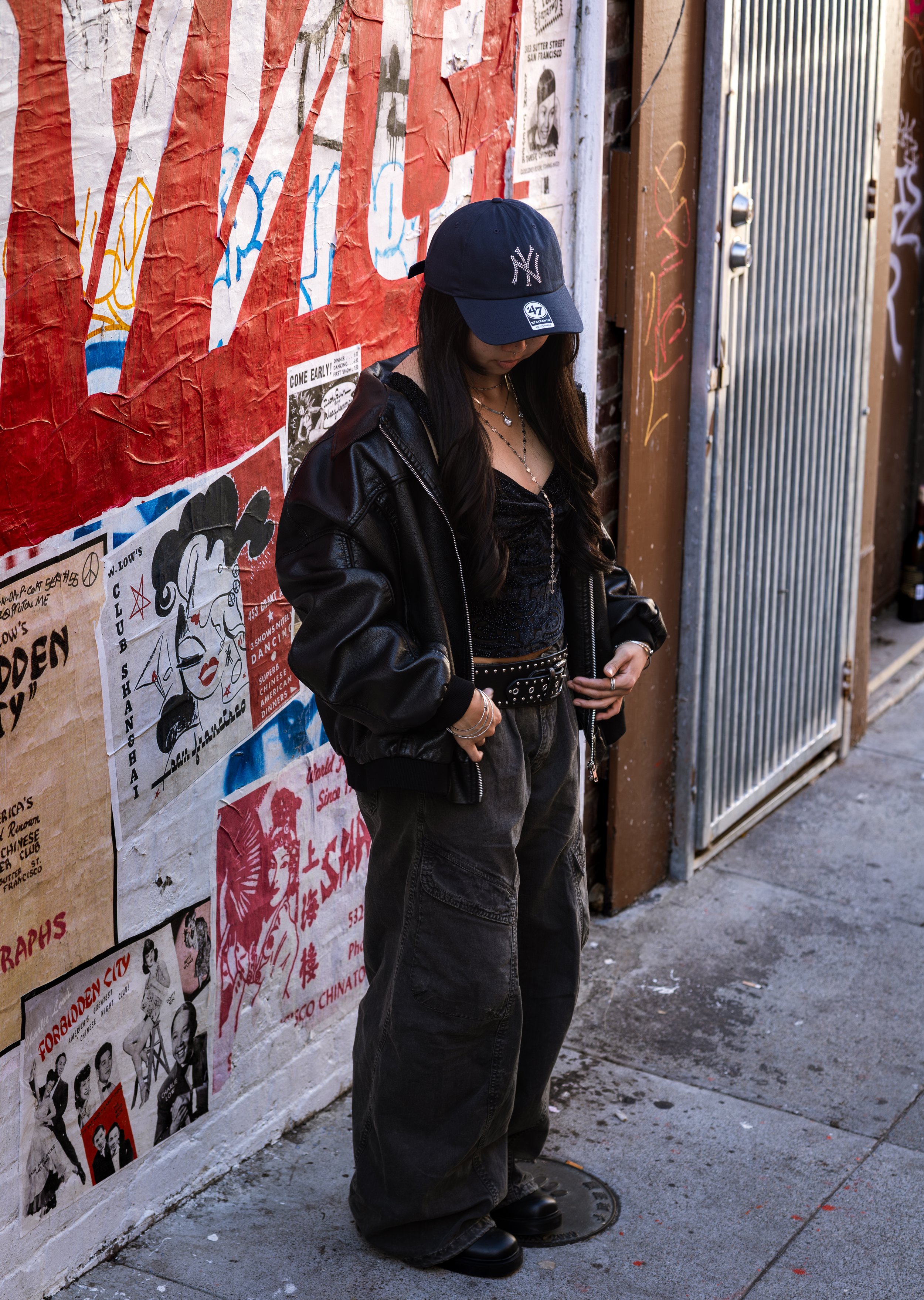 A woman standing on a city sidewalk with her head tilted downward, wearing a black baseball cap with the New York Yankees logo, a black leather jacket, black pants, and black boots. She is outdoors near a wall covered with various colorful posters an