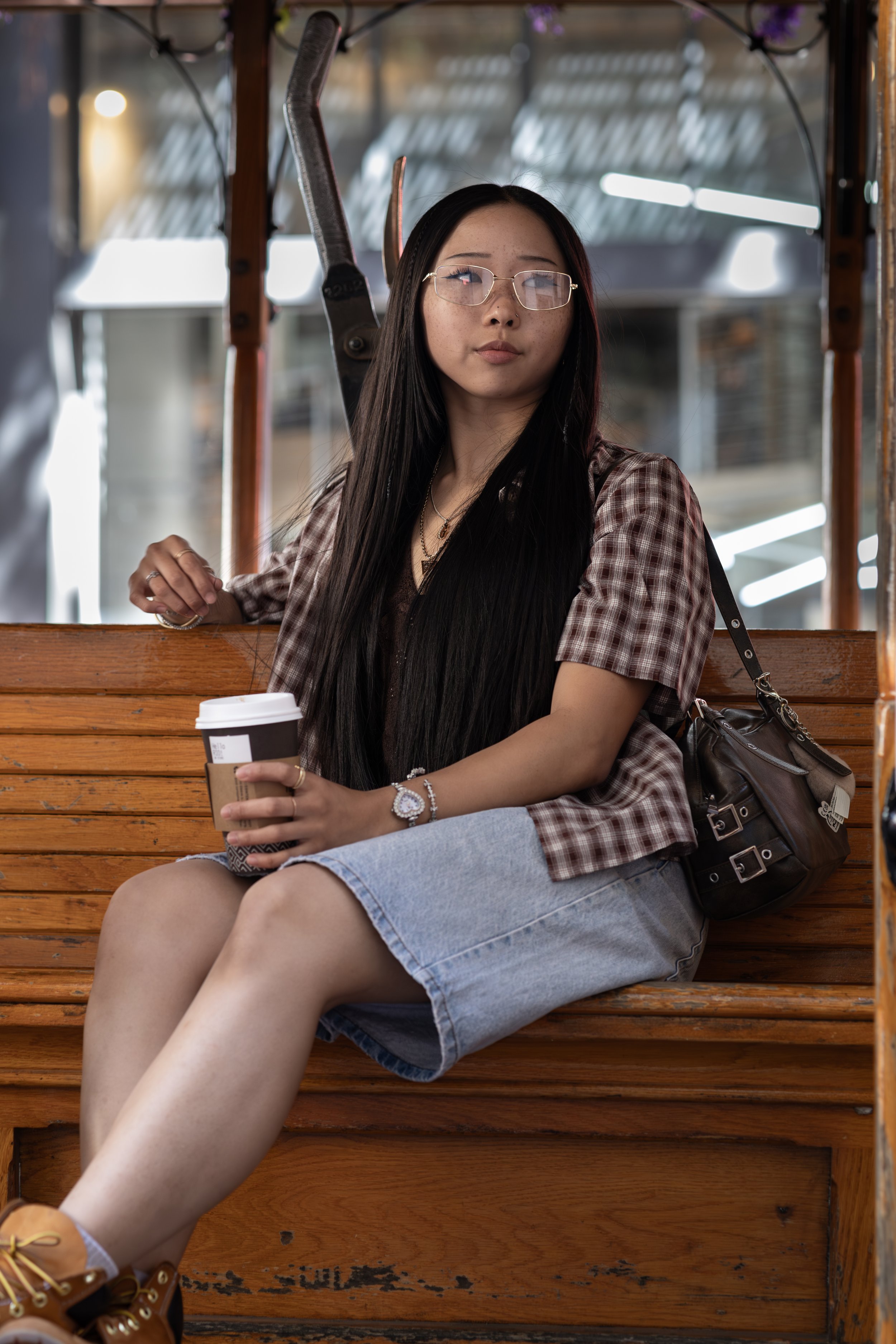 A young woman with long black hair, glasses, and a plaid shirt sitting on a wooden park bench, holding a disposable coffee cup, with a black backpack beside her.