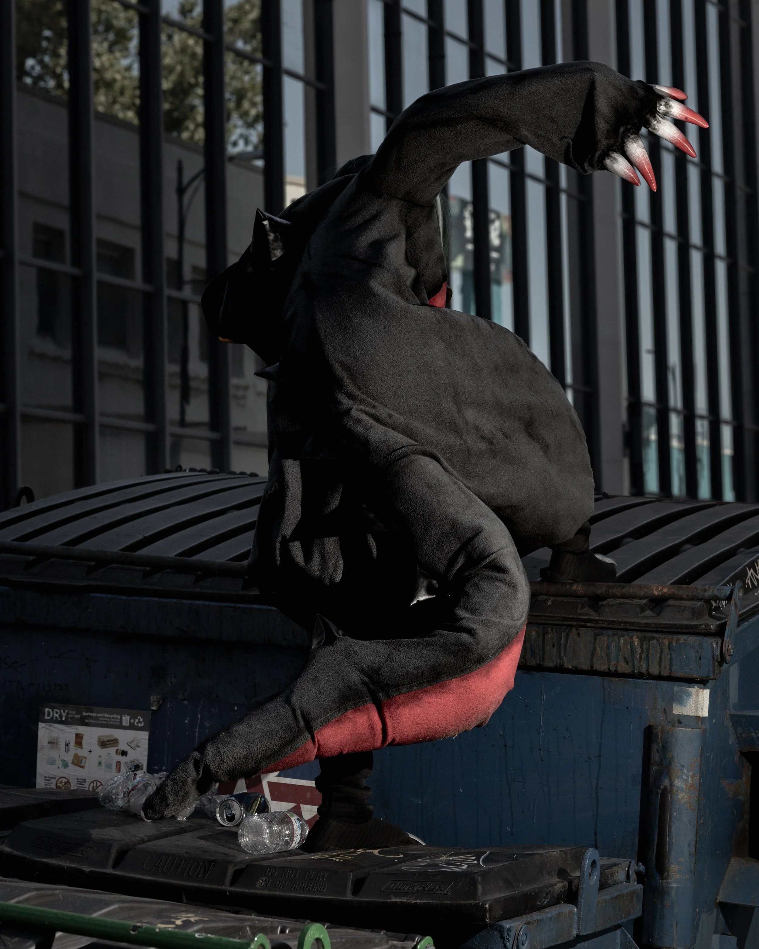 A person wearing black gloves with long red and black acrylic nails, sitting on a bench outside near a metal fence, with trash cans nearby, and water bottles on the ground.