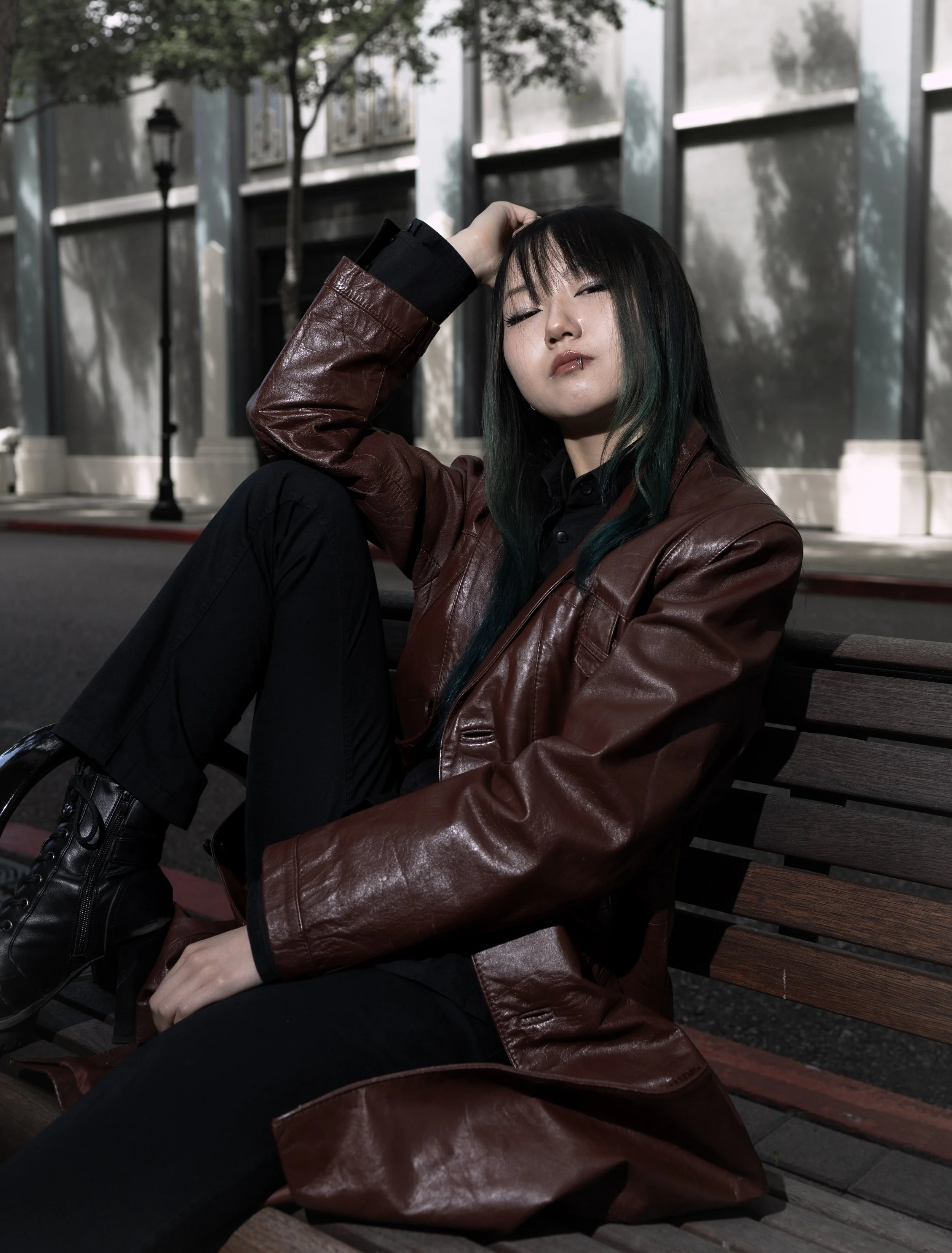 A woman with dark hair and green tips, wearing a brown leather jacket and black pants, sitting on a wooden park bench with her eyes partially closed, resting her head on her hand, outdoors in front of a building with large windows and trees.