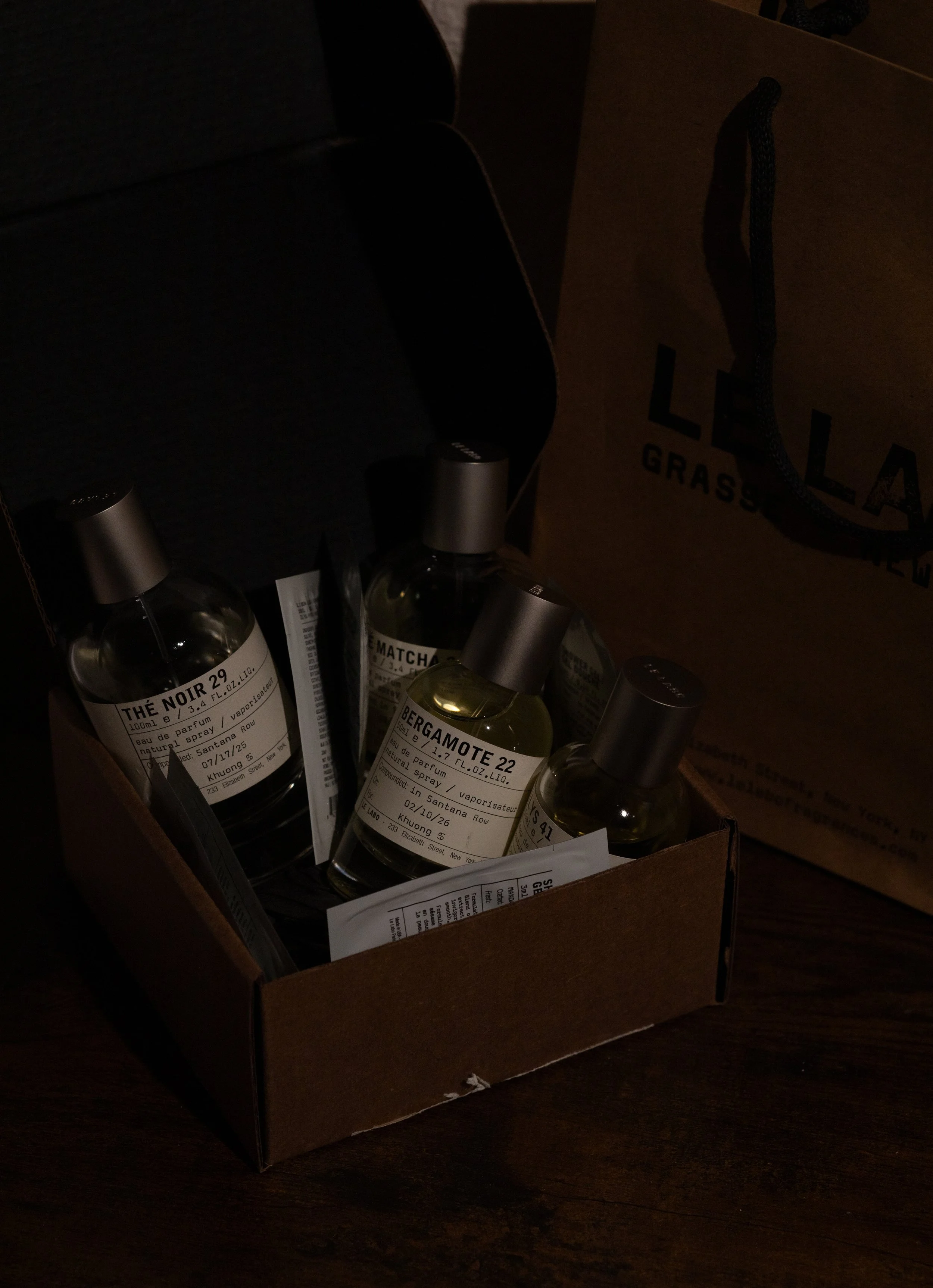 A cardboard box contains several bottles of perfume with white labels and black text. The labels indicate the perfume scents, such as THE NOIR 29 and BERGAMOTE 22. The box appears to be on a dark wooden surface.