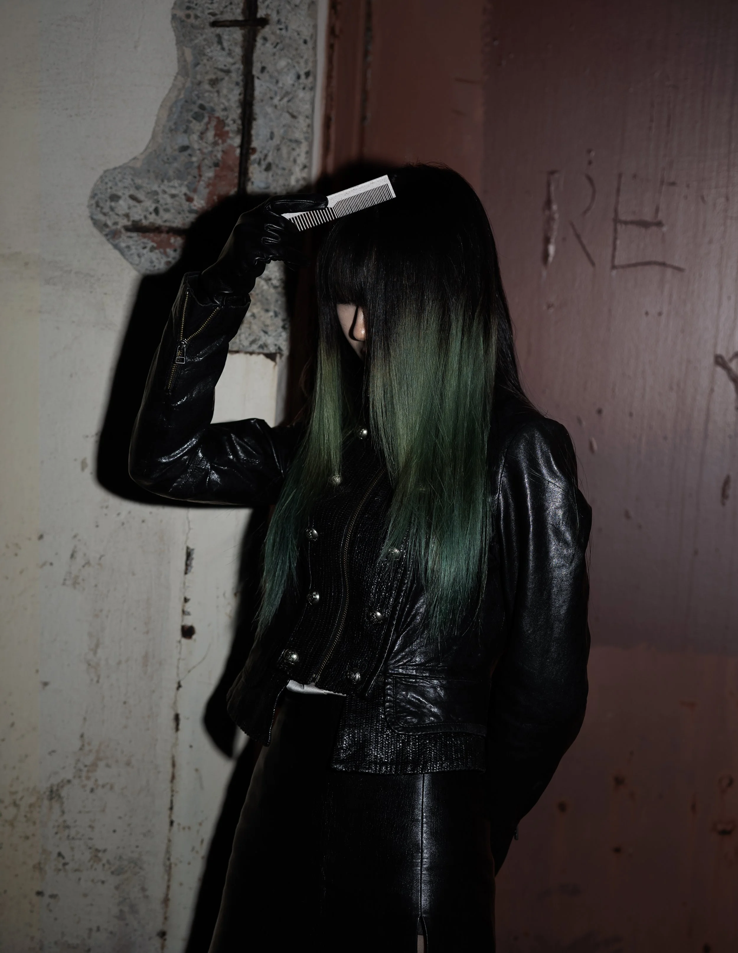 Person with black hair and green tips, wearing a black leather jacket, holding a comb to their hair, standing against a concrete wall with some graffiti.