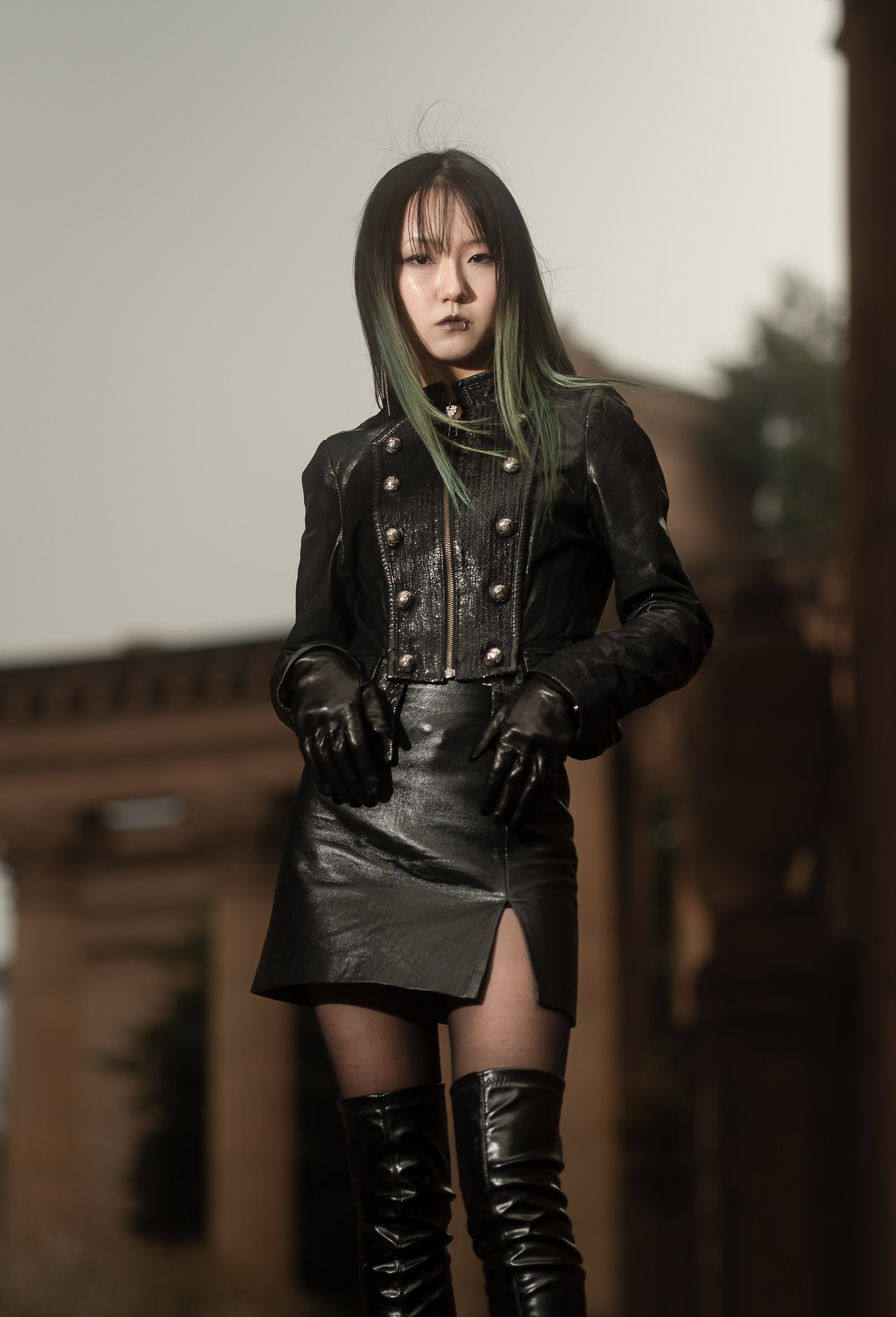 A woman wearing a black leather jacket with metallic buttons, a black leather skirt with a slit, sheer black thigh-high stockings, and black thigh-high boots, stands outdoors in front of a blurred background.