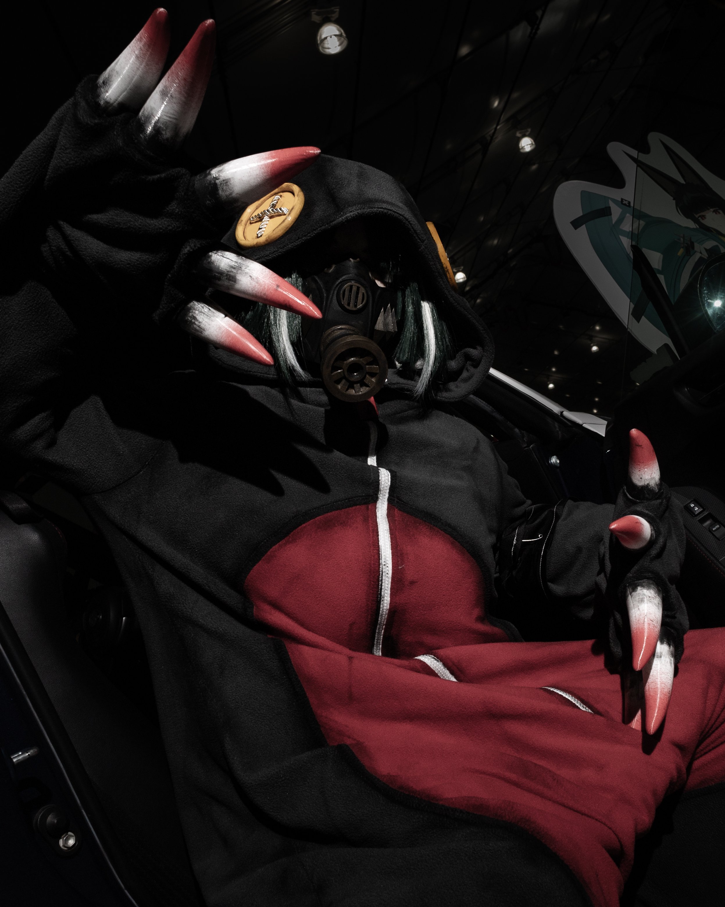 A person wearing a black hoodie with a wooden button and a rope-shaped letter 'X' on it, red and white long, pointed nails, and a red jacket underneath, sitting inside a vehicle with a dark interior and a partial view of a comic-style graphic on the 