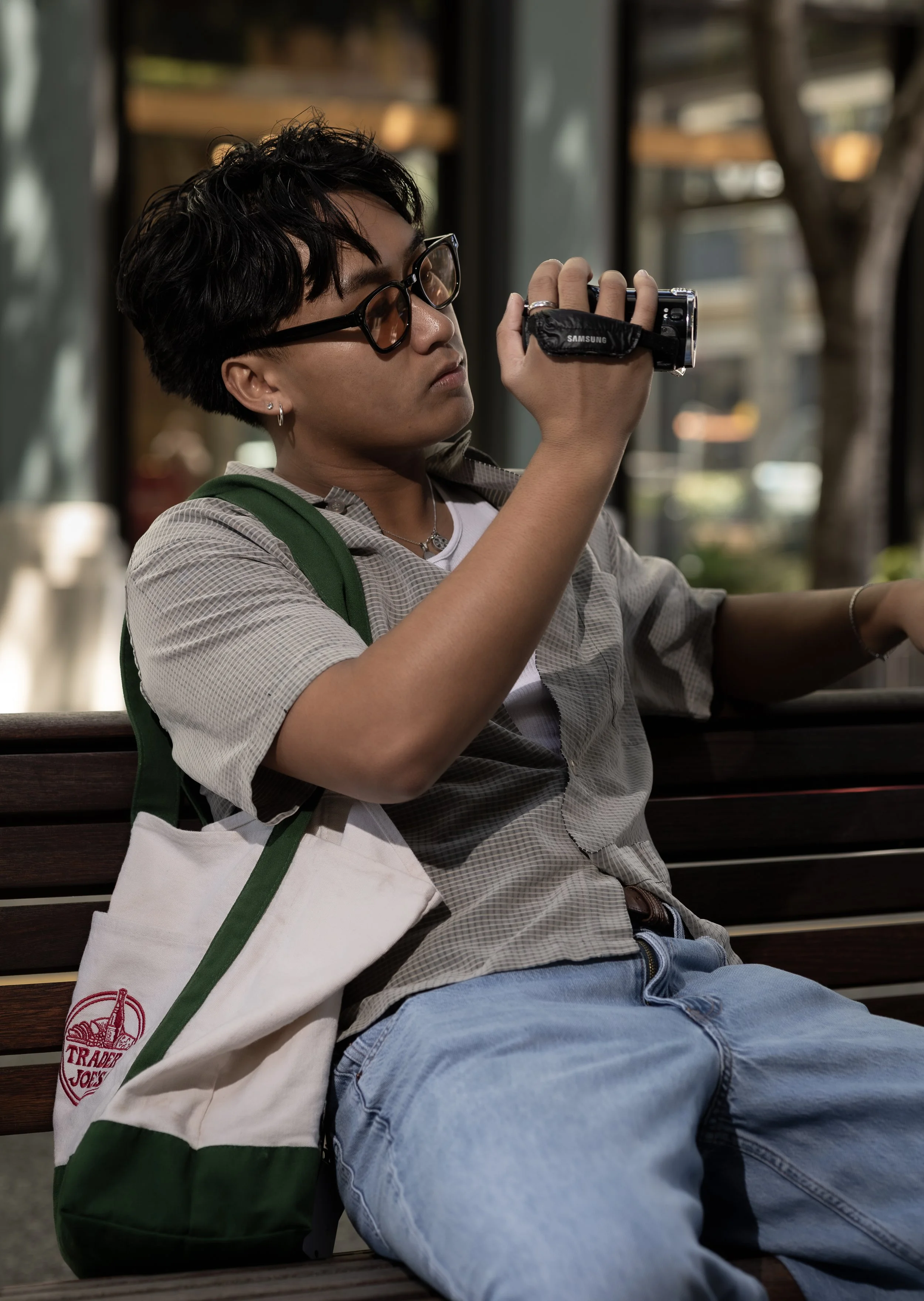 A person sitting on a wooden bench, wearing glasses and a striped shirt, holding and inspecting a small camera. They have a green and white tote bag on their lap and are wearing light blue jeans.