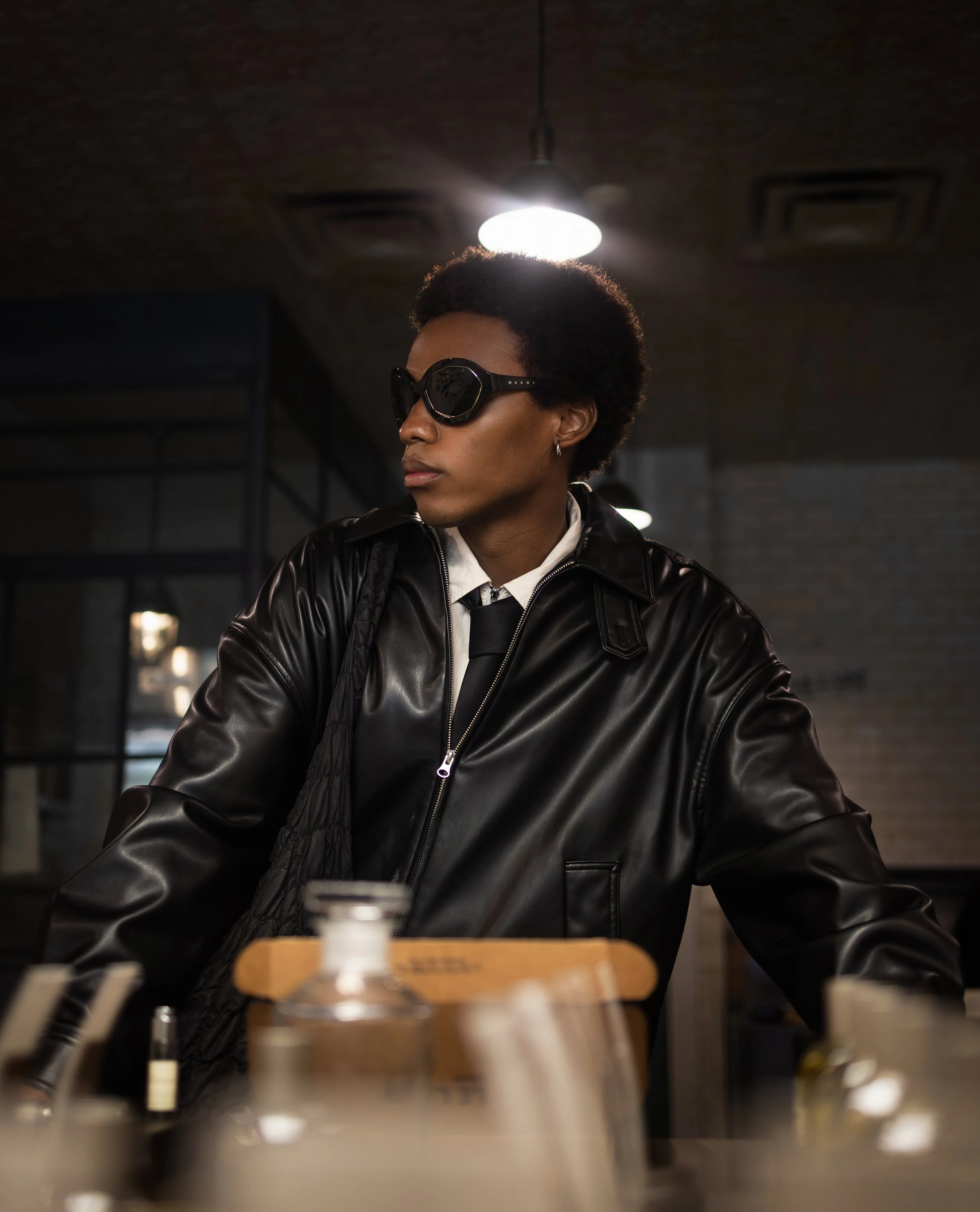 Person wearing black sunglasses and a black leather jacket sitting at a table with bottles in the foreground and a light fixture above in a dimly lit room.