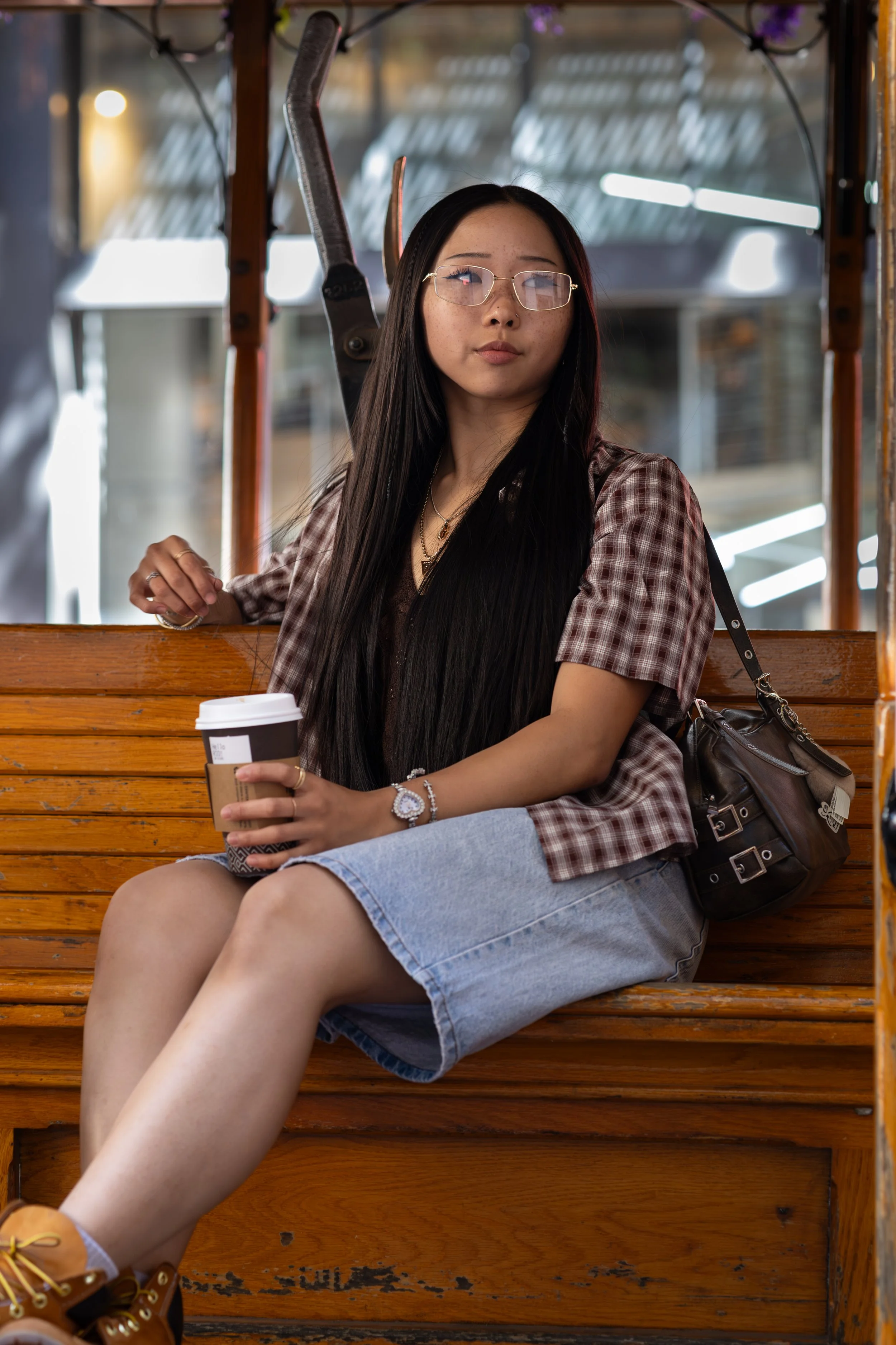 A young woman with long dark hair, wearing glasses, a brown checked shirt, a denim skirt, and yellow sneakers, sitting on a wooden bench in a cafe or restaurant. She holds a to-go coffee cup in her left hand and appears to be looking at the camera wi