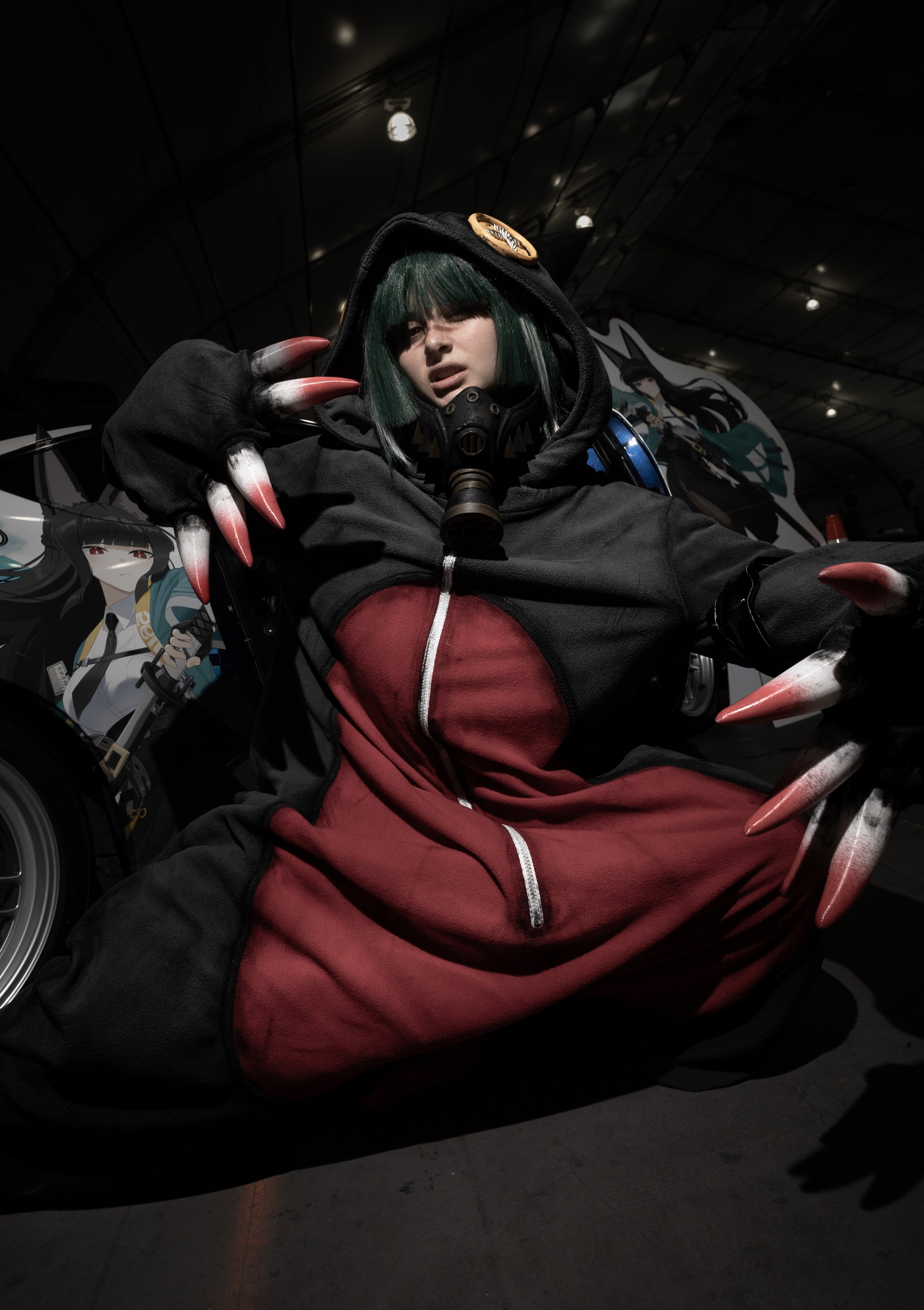 A person dressed as a character with green hair, wearing a black hoodie and red pants, sitting with claws on their hands, in a dark indoor setting with anime and fantasy themed decorations in the background.