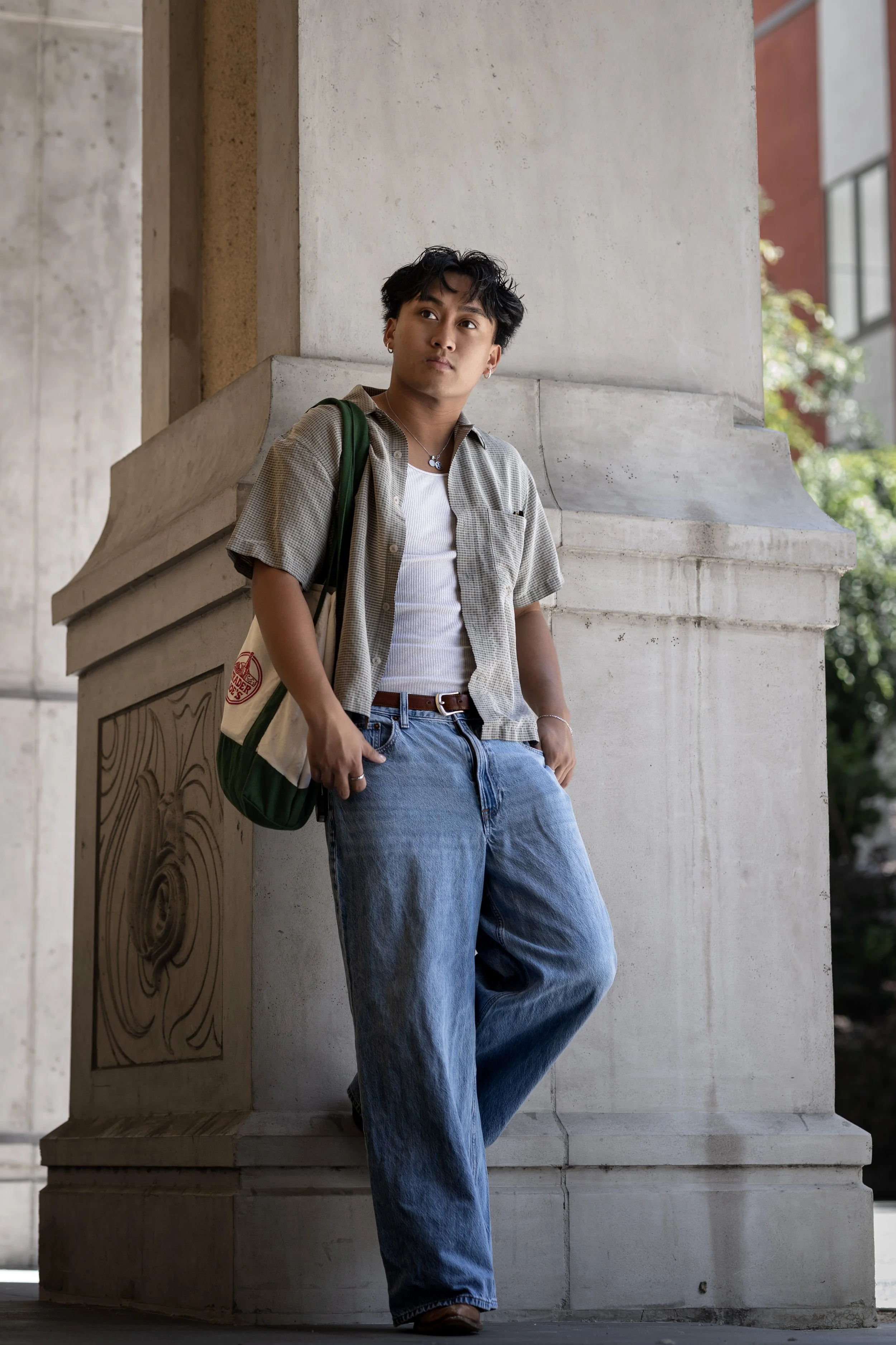 A young man with dark, wavy hair leaning against a stone column with architectural carvings, wearing a checkered shirt over a white tank top, blue jeans, and a green bag on his shoulder, outdoors on a sunny day.