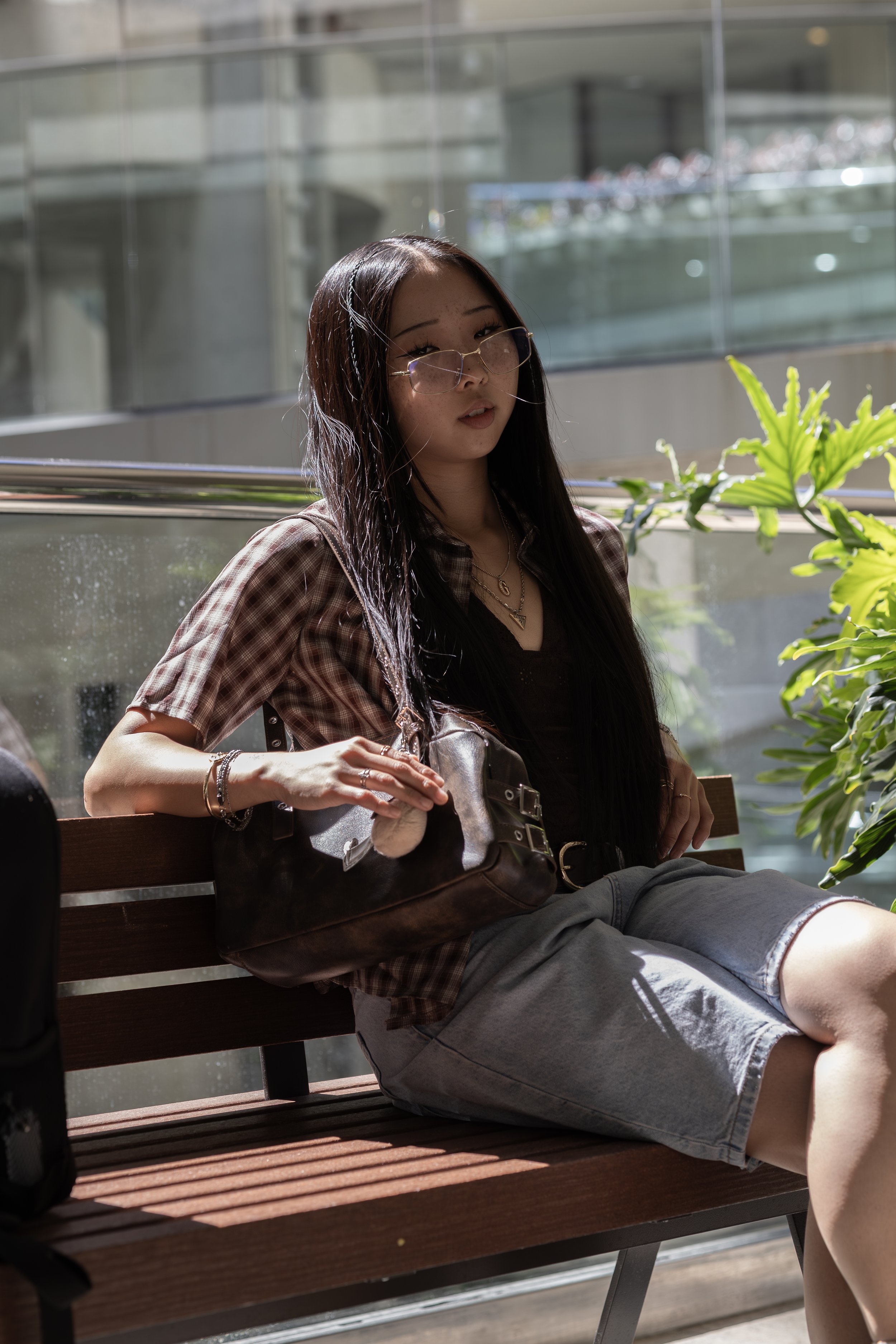 Young woman with long dark hair, wearing glasses, a checkered shirt, and shorts, sitting on a wooden bench in a modern indoor space with glass walls and green plants, holding a brown bag.