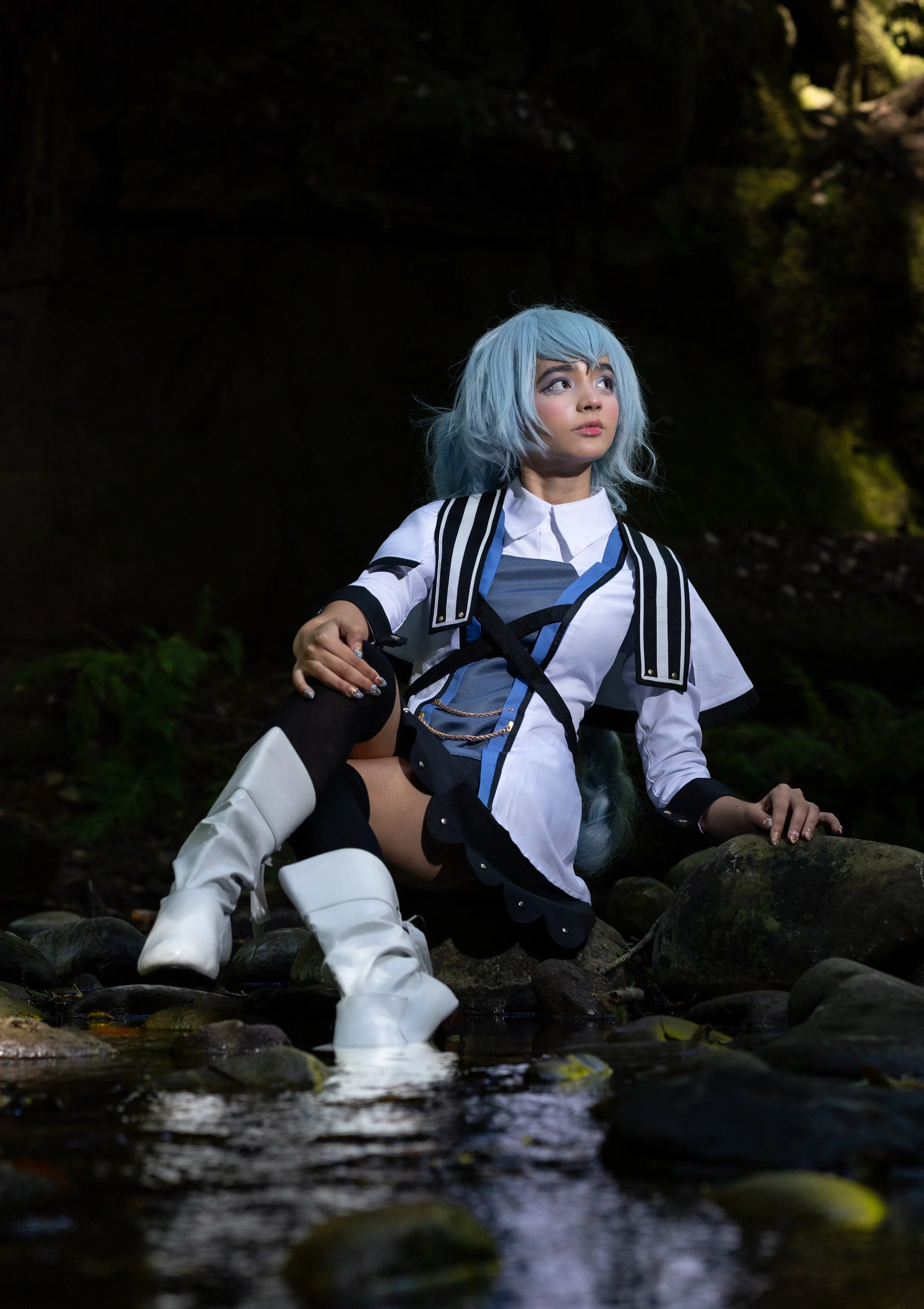 A person with light blue hair, wearing a white, black, and blue costume, sitting on rocks near a stream in a forested area, looking upward with a contemplative expression.