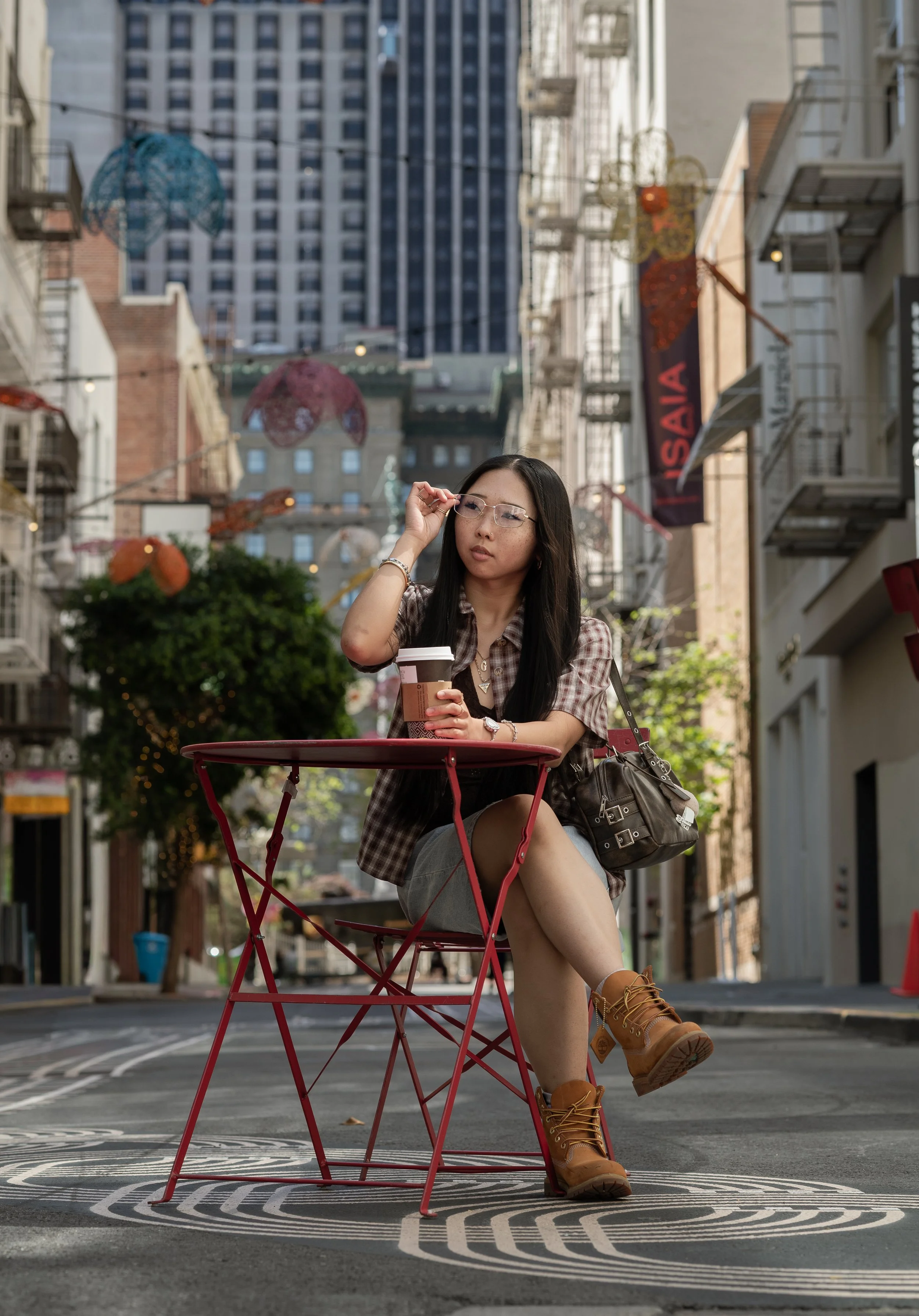 A young woman sitting at a small red outdoor table in an urban street, holding a coffee cup and adjusting her glasses. She is wearing tan boots, a checkered shirt, and has a black handbag hanging on her side. The street features colorful hanging deco