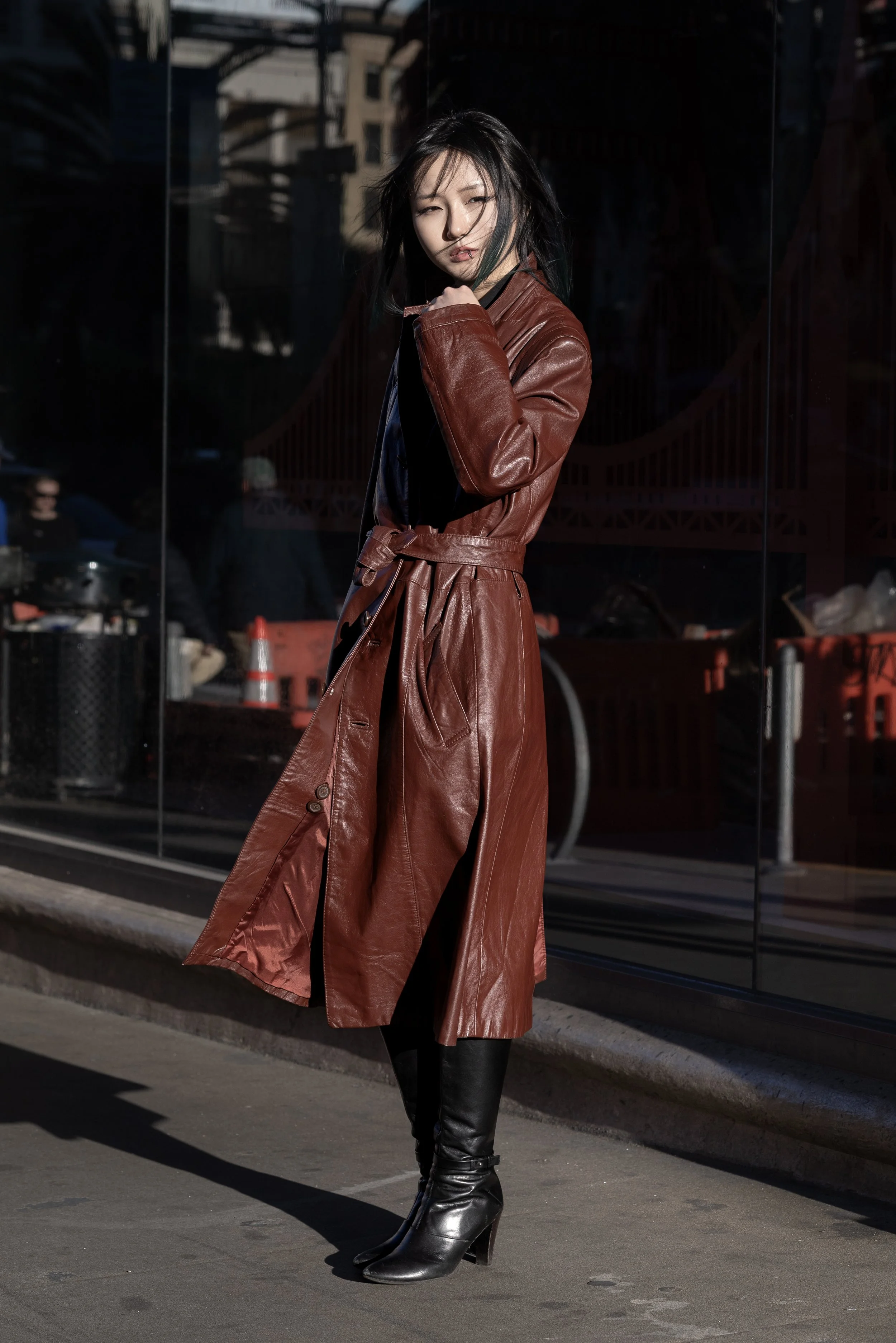A woman in a long brown leather trench coat and black high-heeled boots standing outside on a city street, with her hair blowing in the wind. Reflection of buildings in the background glass.