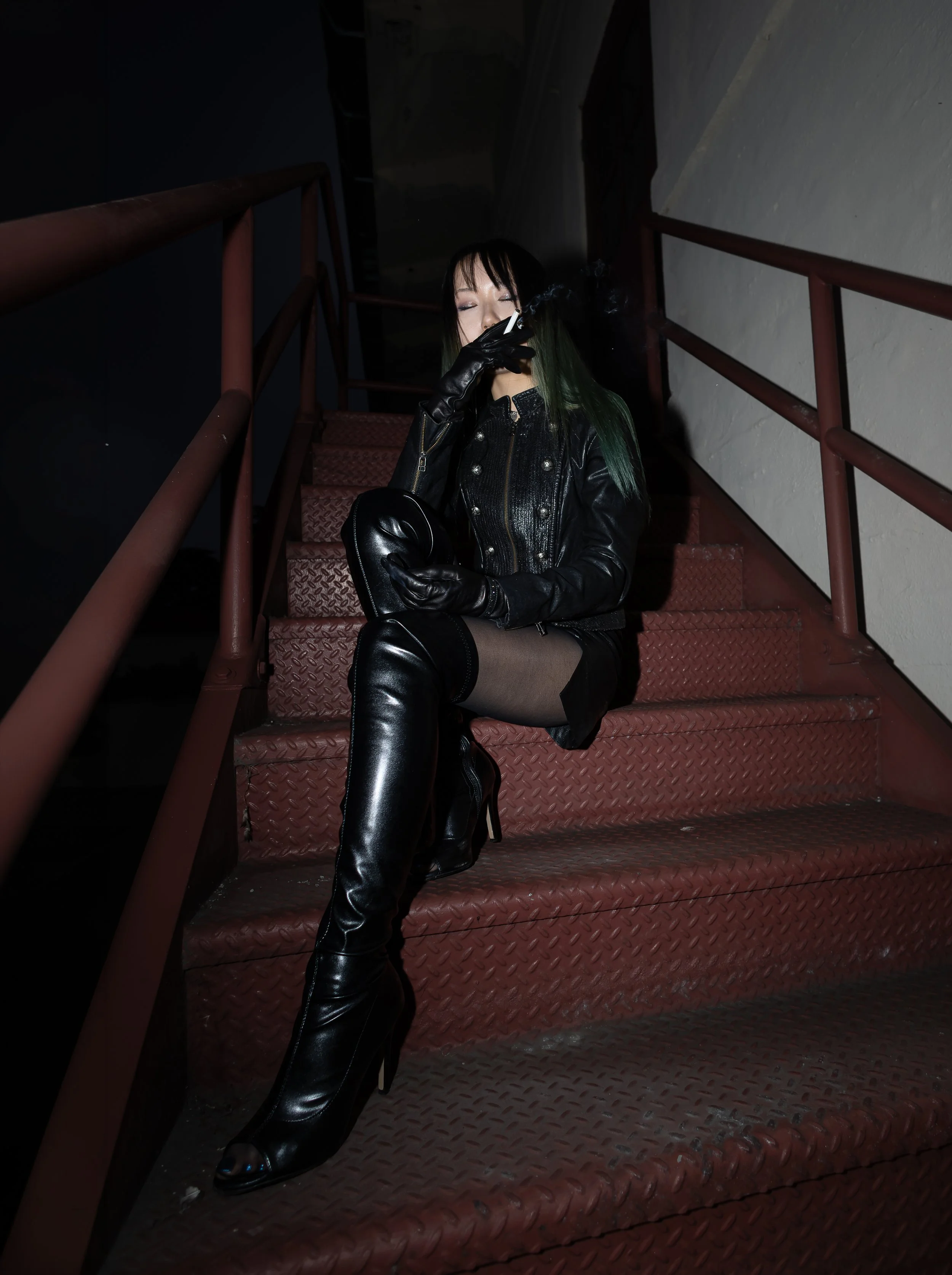 Woman with dark hair and green tips sitting on a staircase, wearing black leather gloves, a leather jacket, and thigh-high black leather boots, smoking a cigarette in a dimly lit setting.