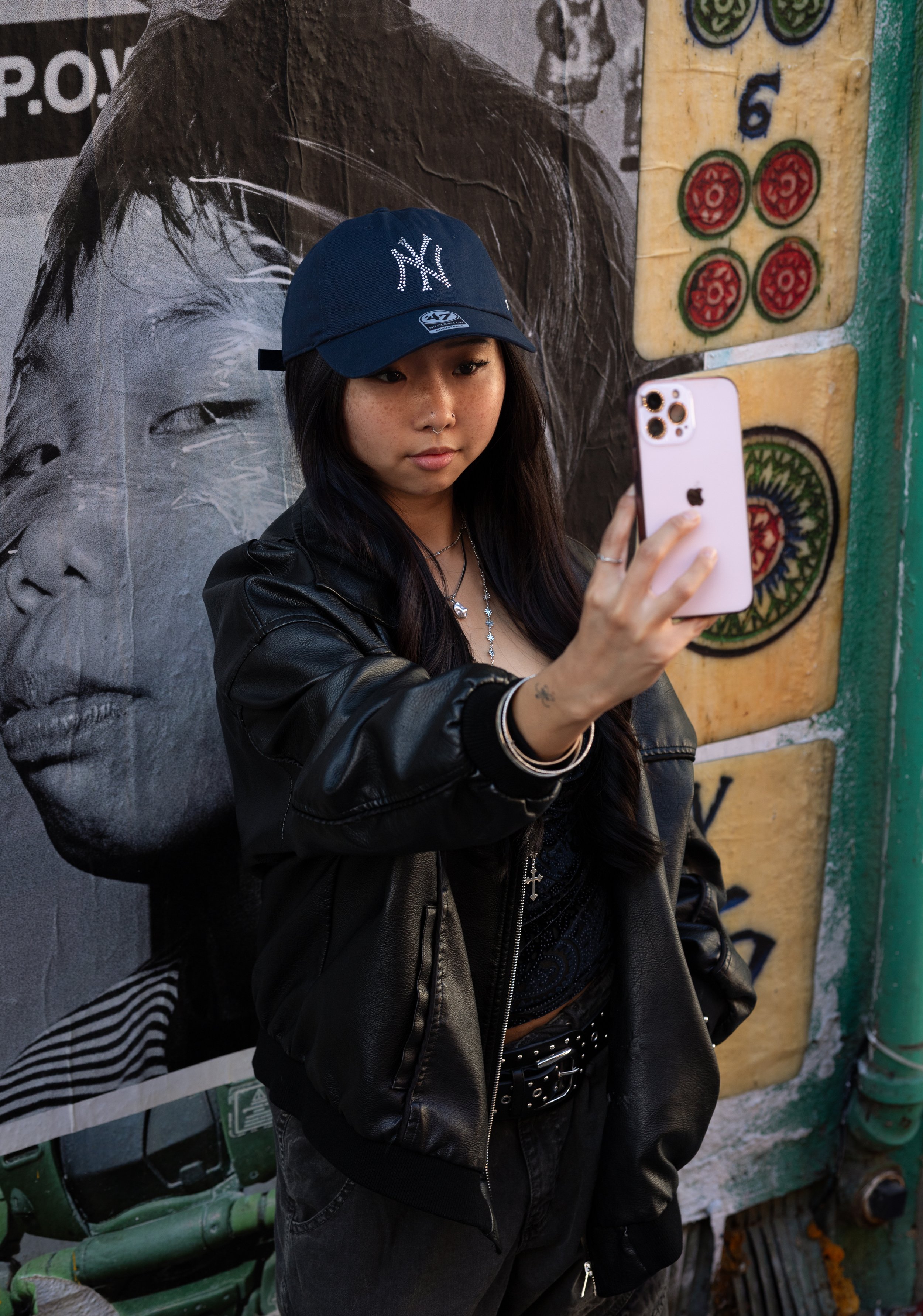 A young woman with dark hair and freckles taking a selfie with a smartphone. She is wearing a navy blue cap with the Yankees logo, a black leather jacket, a black top, and a necklace. She is standing in front of a mural featuring a grayscale face and