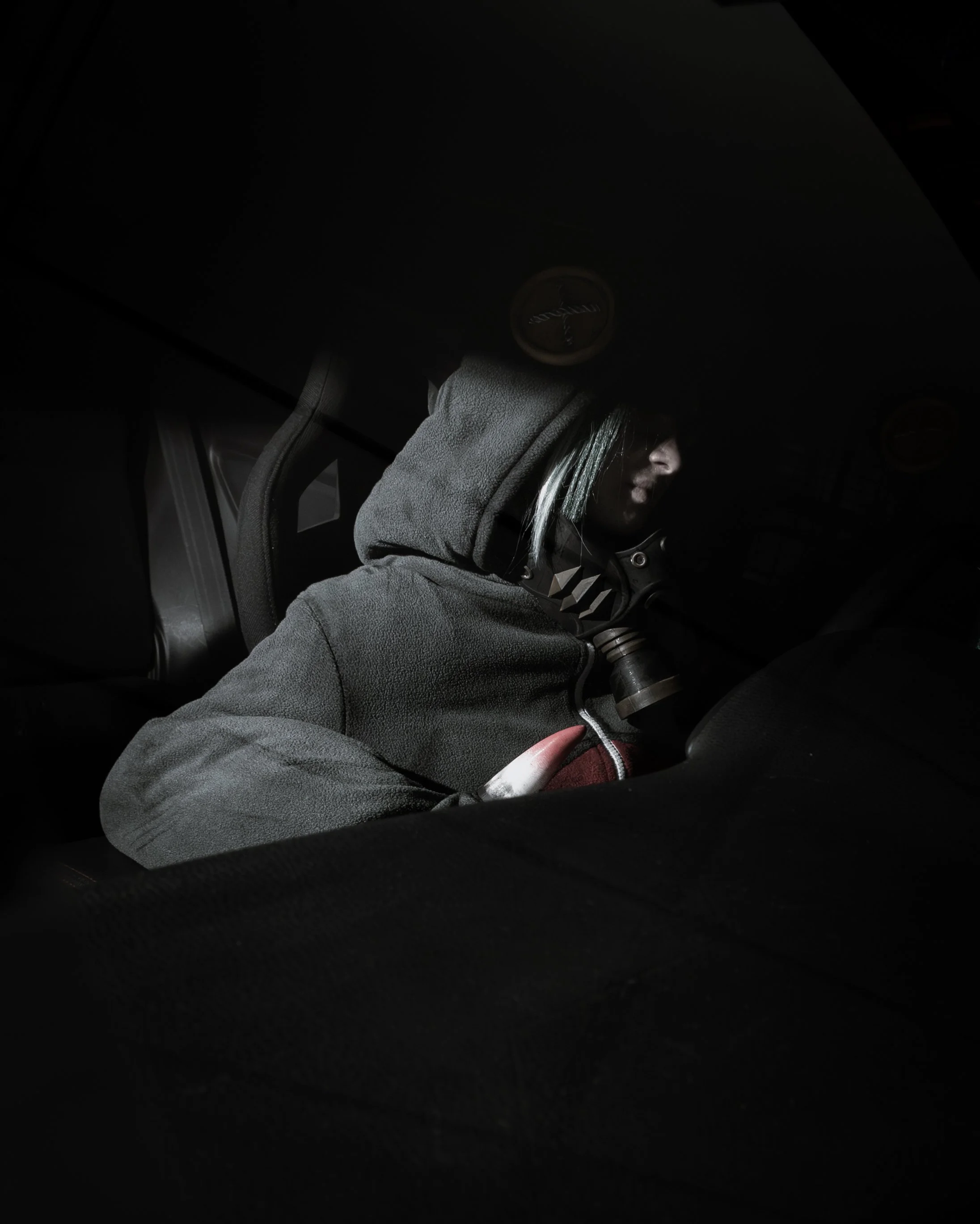 A person wearing a grey hoodie with white hair partially visible, sitting on a vehicle with a dark interior, possibly a car, with a very dark environment around.