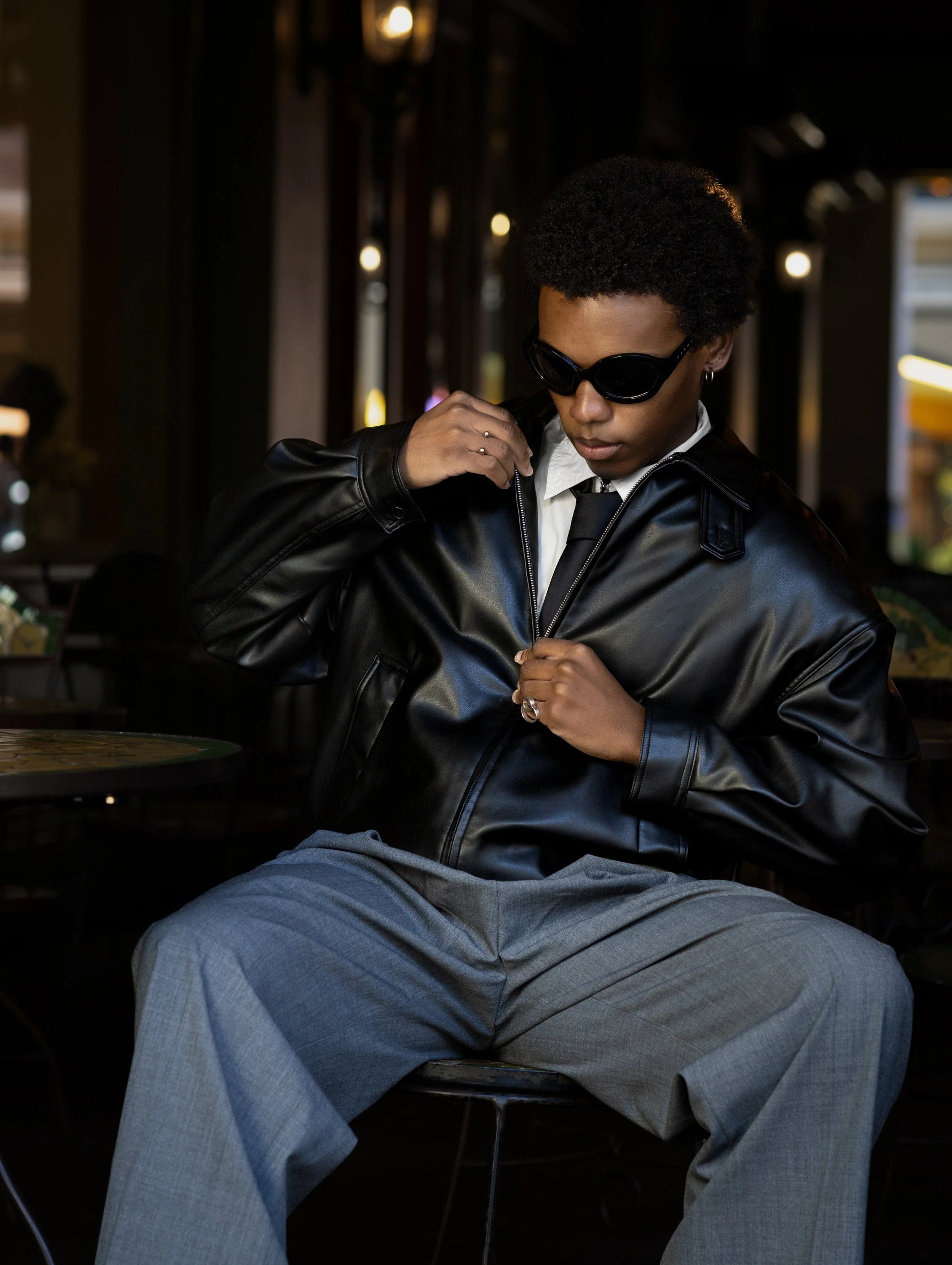 A person with dark skin, short curly hair, wearing sunglasses, a black leather jacket, white shirt, and black tie, sitting indoors with a dark background and some blurred lights behind them.