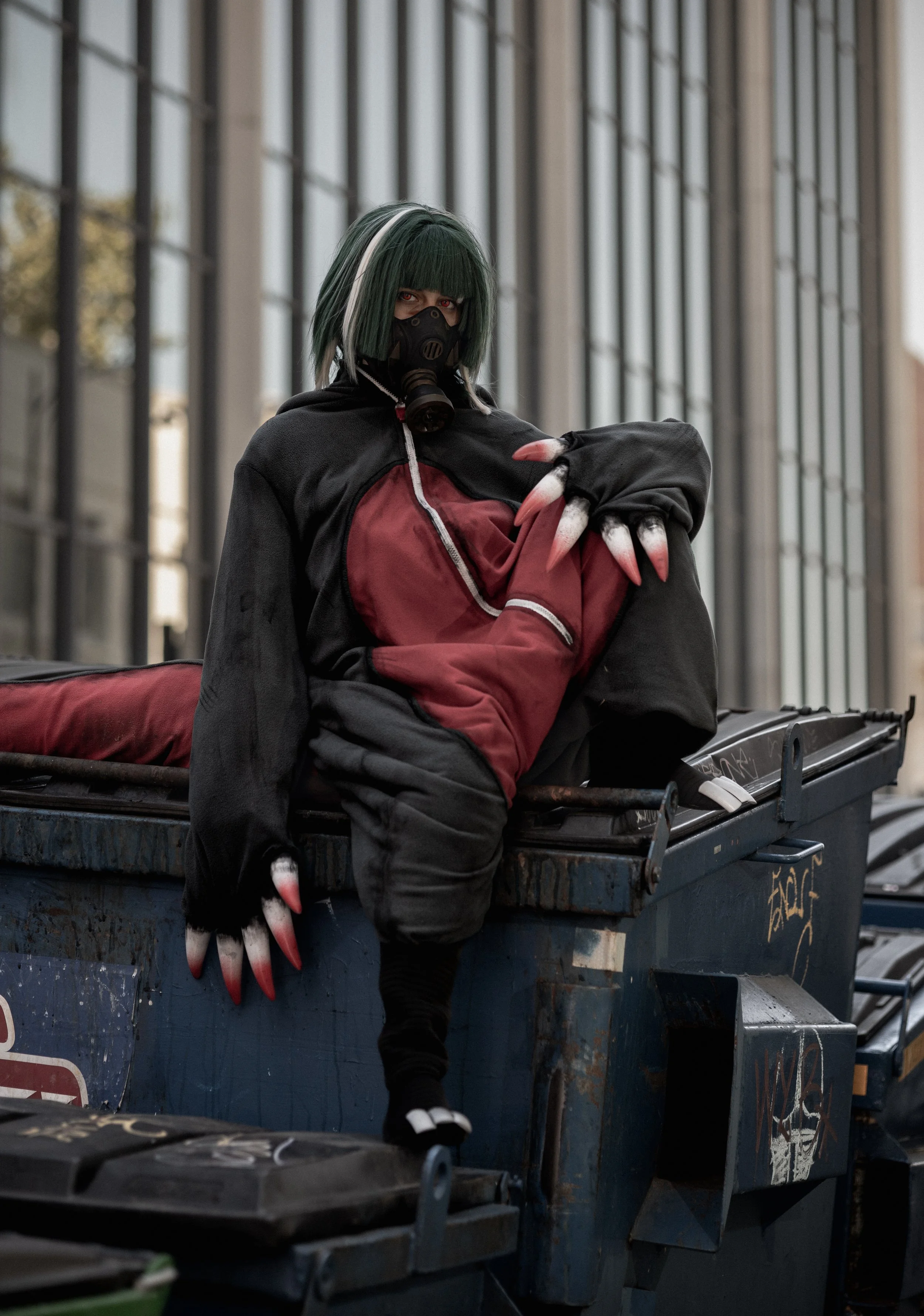 Person dressed in dark clothing with a gas mask, green and white wig, and clawed gloves, sitting on a dumpster in front of a modern glass building.