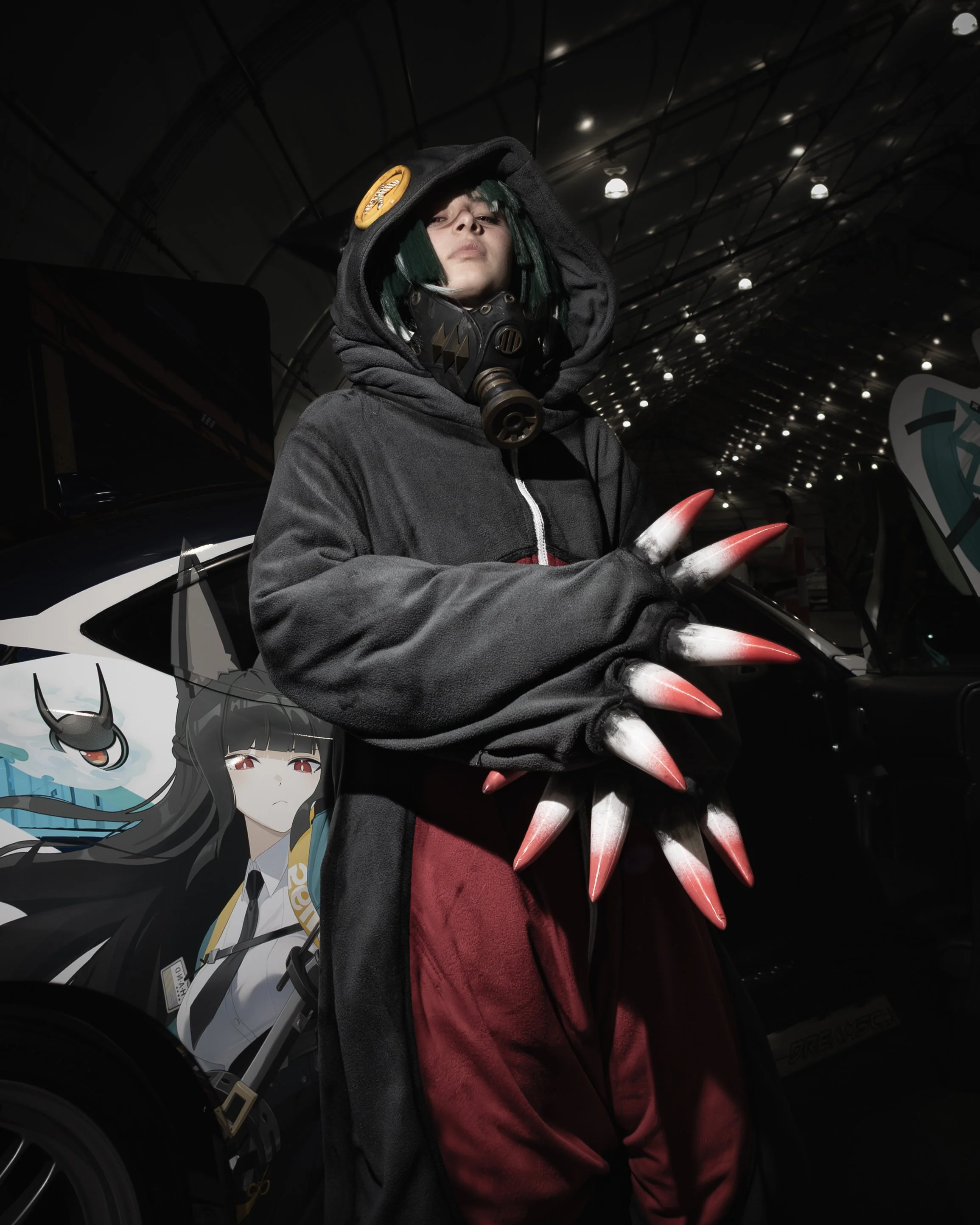 Person dressed in a black hoodie with green hair, wearing a gas mask, with large red claws, standing in front of a black vehicle featuring anime-style artwork of a girl with black hair and red eyes.