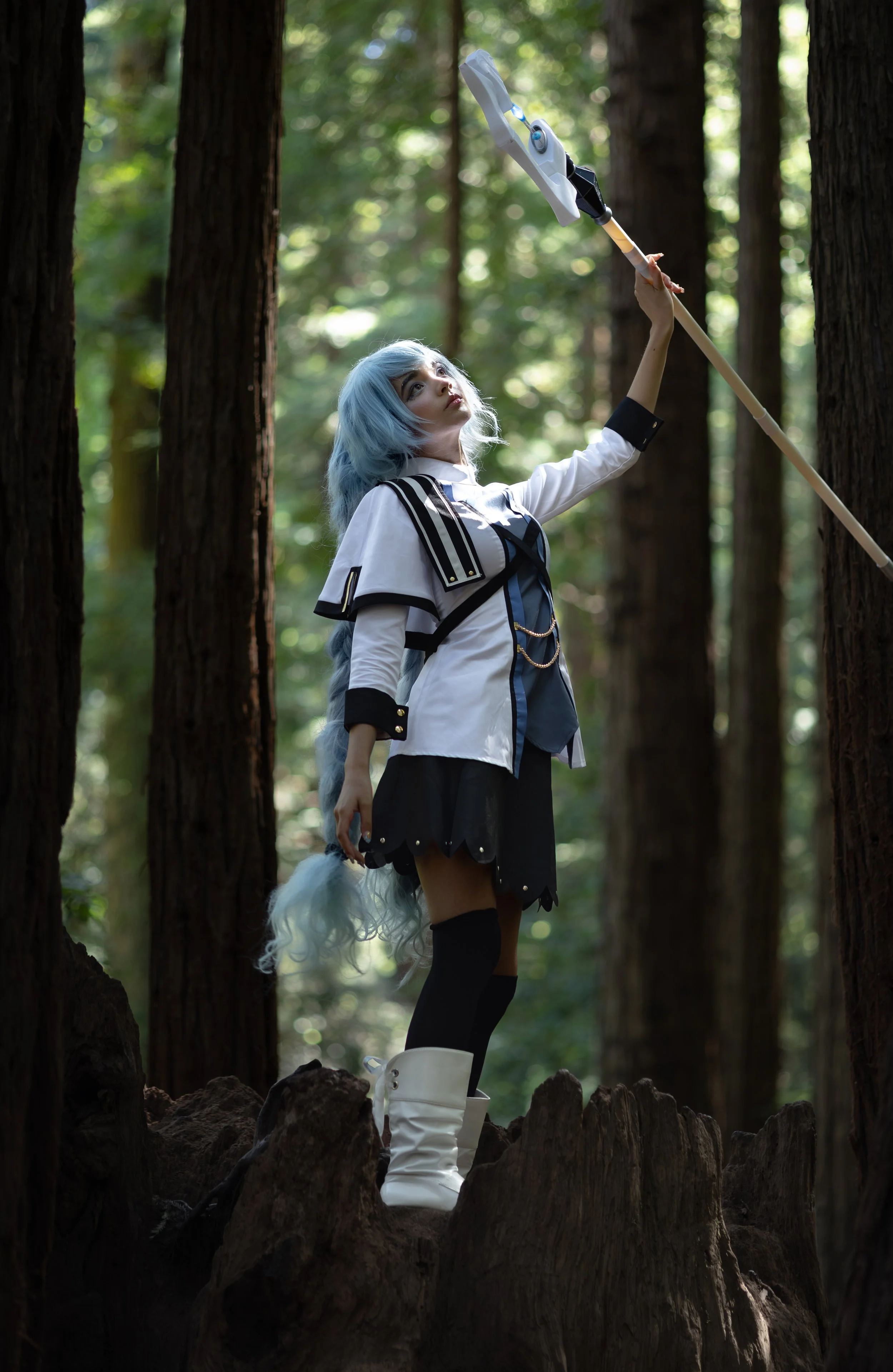 A person with long, light blue hair and a white tail stands in a forest with tall trees, holding a long staff aimed upward. They are wearing a costume with a white and black color scheme, knee-high black socks, and white boots.