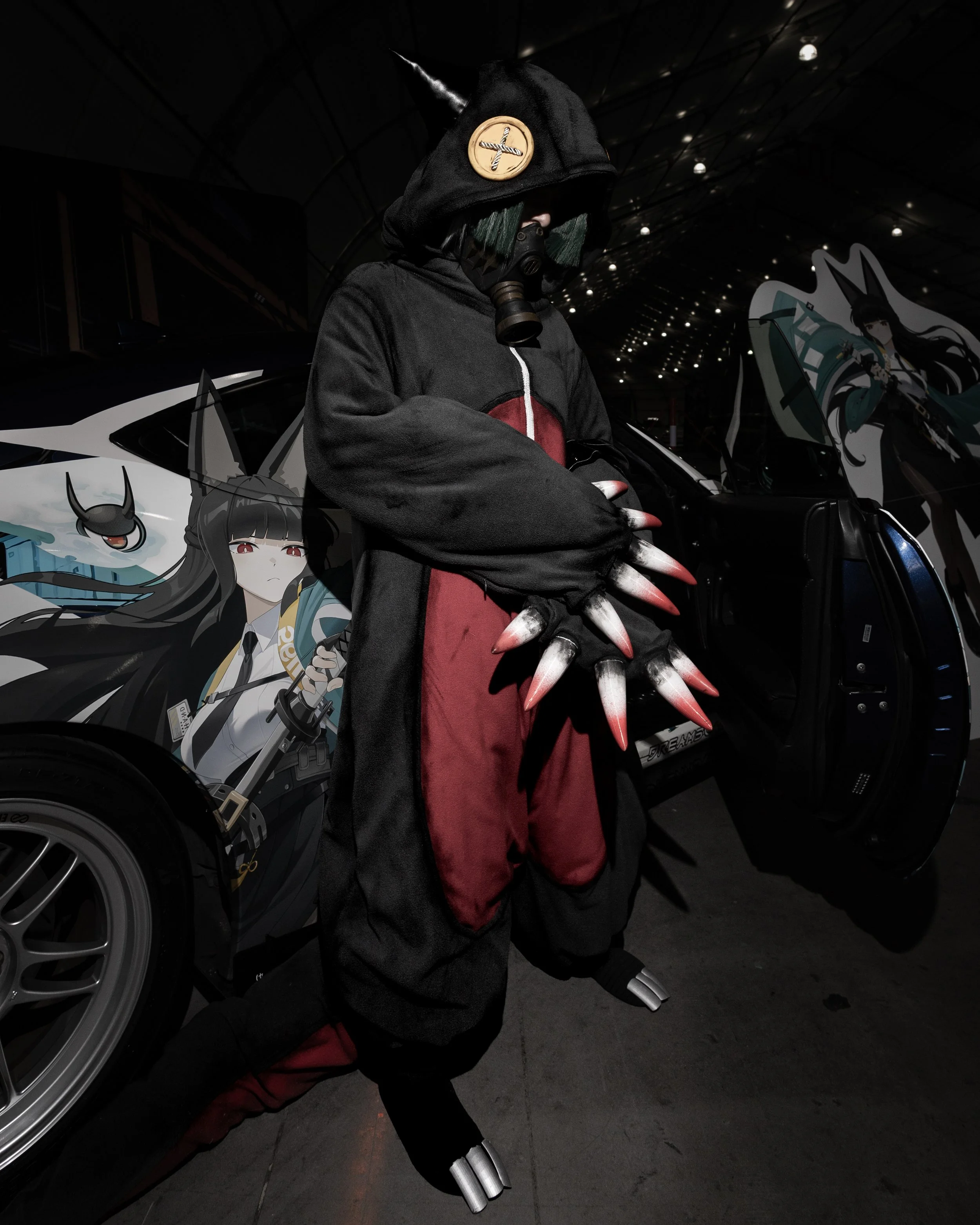 Person in cosplay costume with a hood, gas mask, and claws, standing next to a car with anime-style artwork, in a dark indoor setting with ceiling lights.