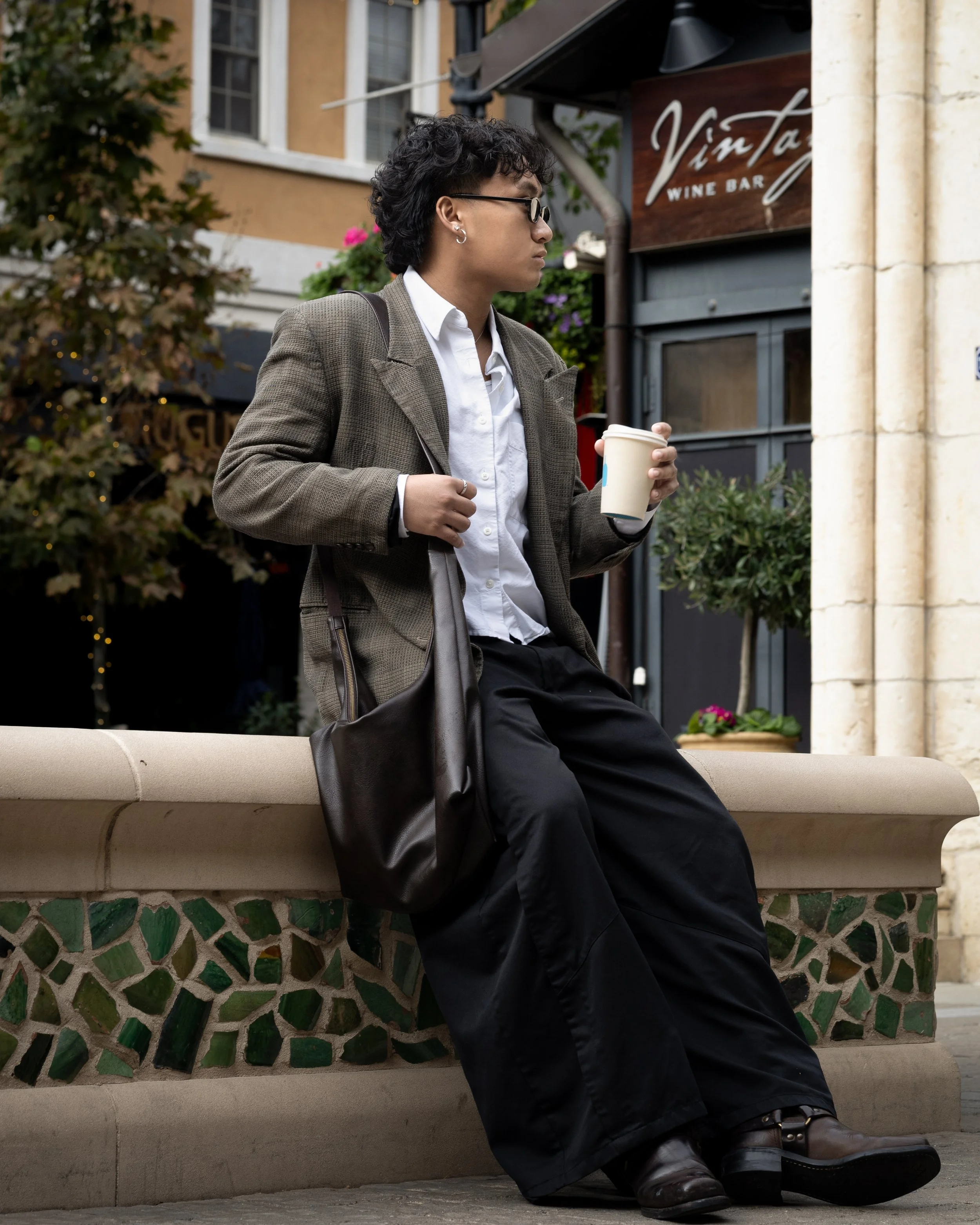 A person with short curly black hair, glasses, a white shirt, blazer, and dark pants sitting on a stone bench, holding a coffee cup, outdoors near a building with a restaurant sign that reads 'VimTo'.