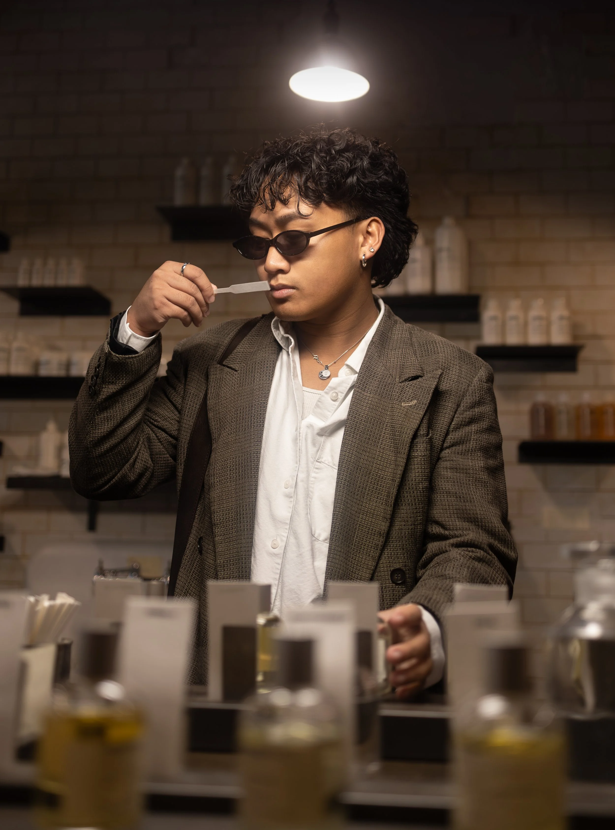 A man with dark curly hair, wearing dark sunglasses, a brown plaid blazer, and a white shirt, is sniffing a sample in a laboratory or pharmacy setting. The background has shelves with bottles and jars, and the foreground shows a row of bottles.