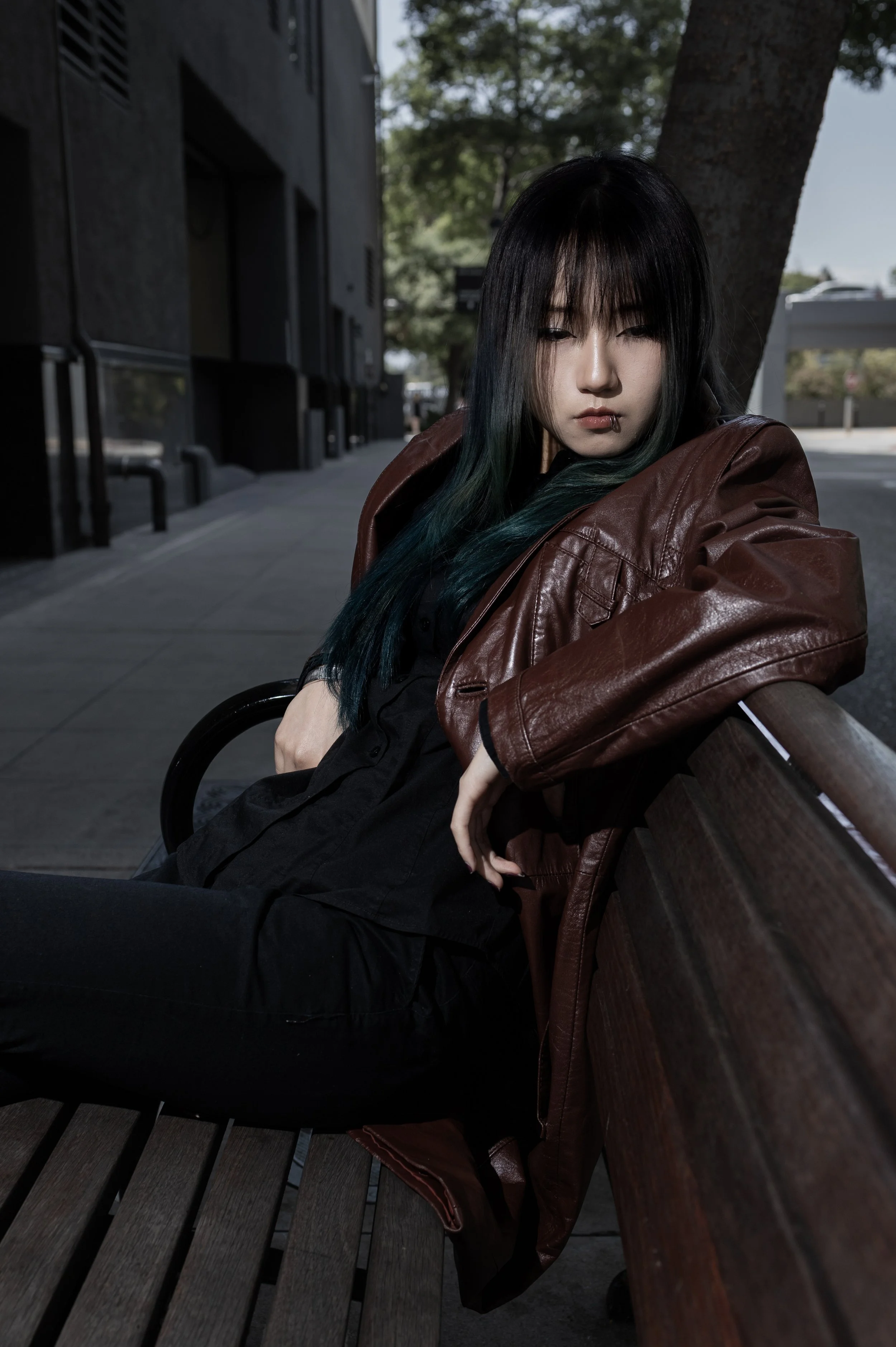A young woman with black hair and subtle makeup sitting on a wooden park bench. She has a metal lip ring and is wearing a brown leather jacket over a black shirt. The setting appears to be outdoors on a sunny day with trees and a building in the back